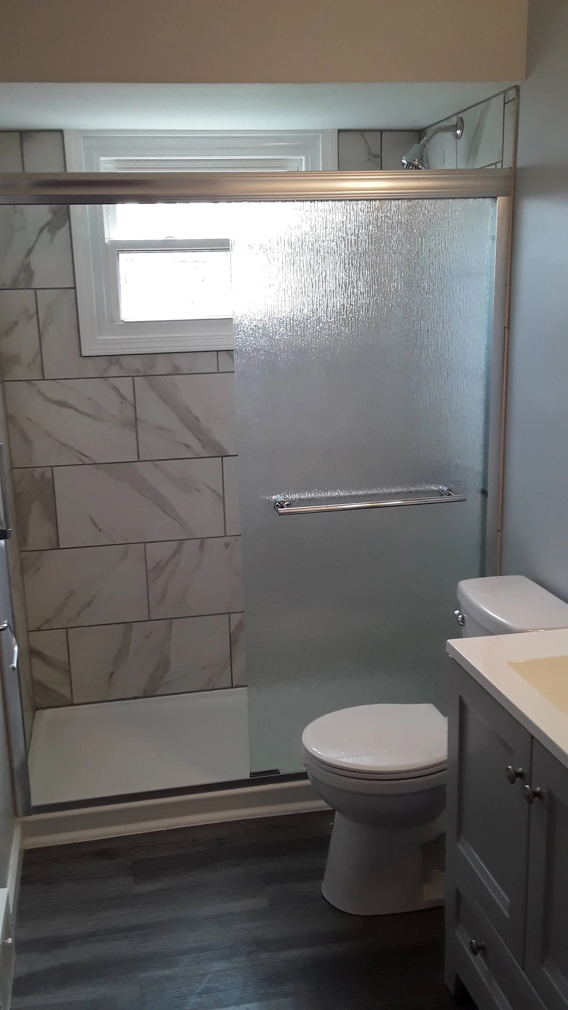Modern bathroom renovation showing custom marble-patterned tile walk-in shower with a low-maintenance glass panel enclosure and updated fixtures.