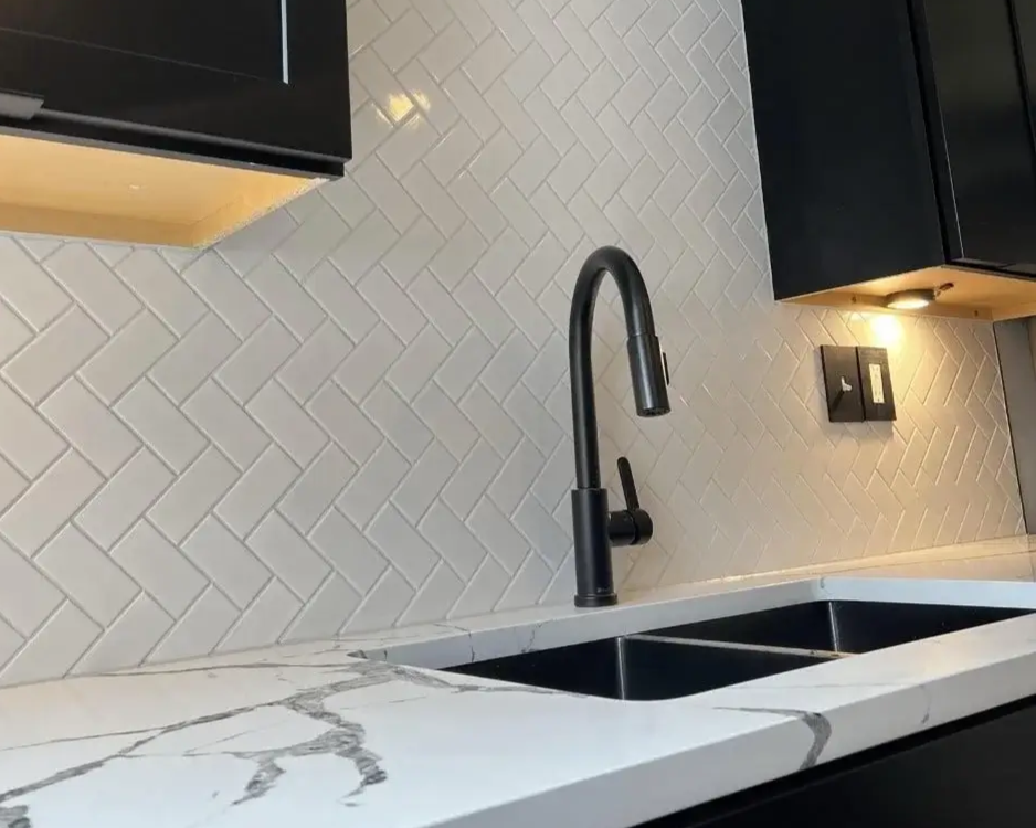 Close-up of kitchen sink with black faucet, white marble countertop with gray veins, beige herringbone tile backsplash, black upper cabinets, and a wall outlet with a light above it.