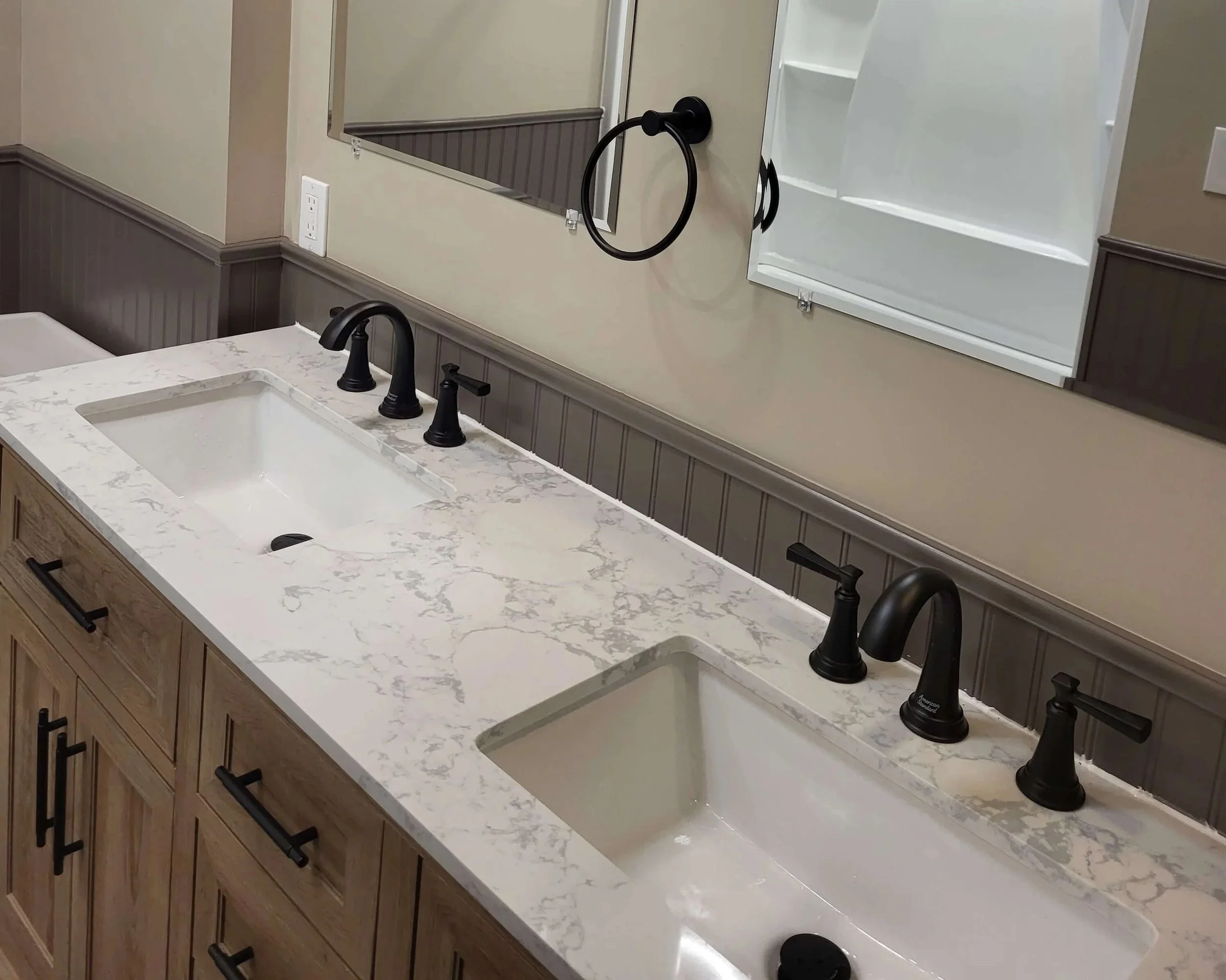 Bathroom vanity with a white marble countertop, two black faucets, a mirror, a wall-mounted black towel ring, and light-colored walls with dark wainscoting.