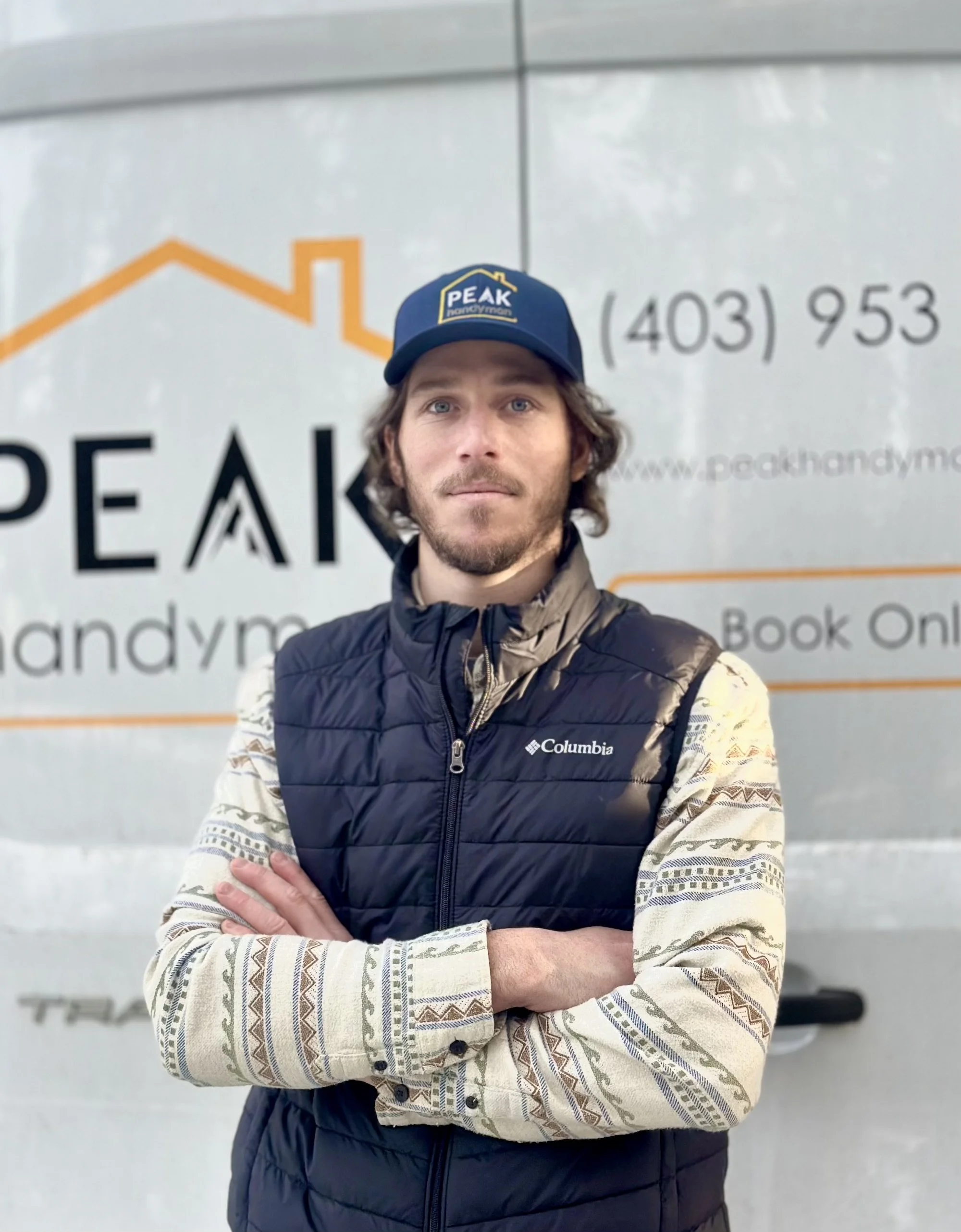 Robin Colas, owner of Peak Handyman Services standing with arms crossed in front of a vehicle with the word PEAK Handyman Services and a phone number on the side.