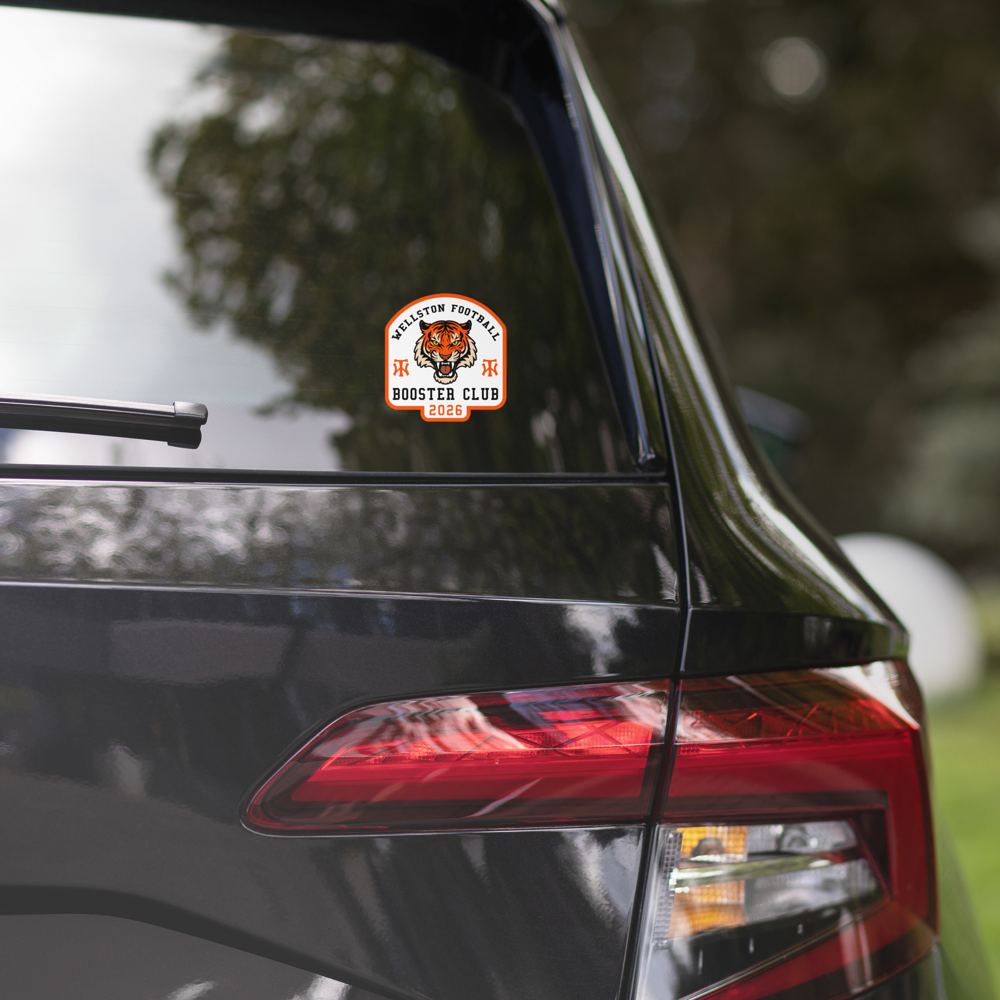A close-up of the rear window and tail light of a black car with a sticker that reads "Wellston Football Booster Club 2026" featuring a tiger mascot.