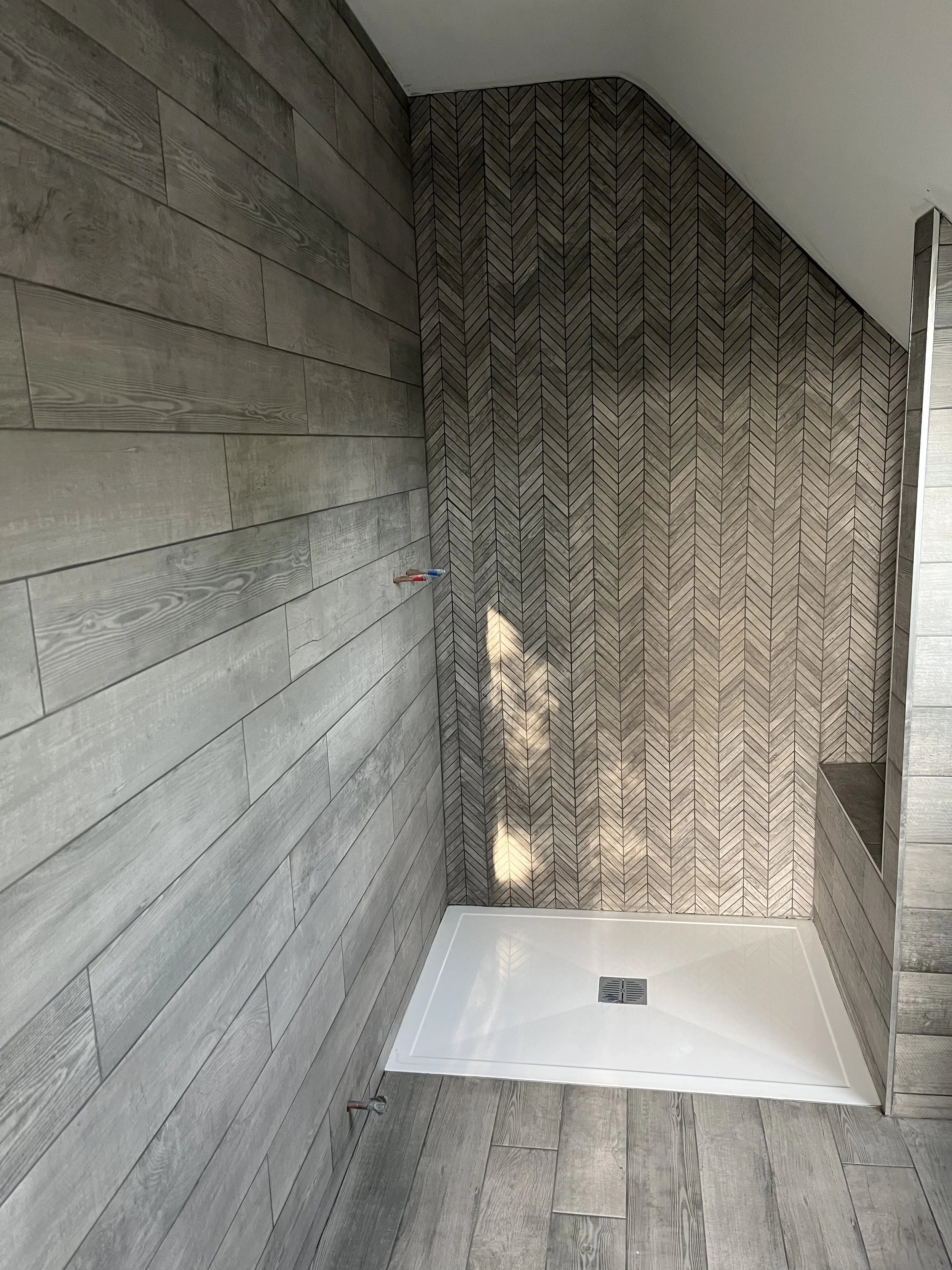 Indoor shower area with a white shower pan, gray textured wood-look tiles on the walls and floor, and a decorative herringbone-patterned accent wall.