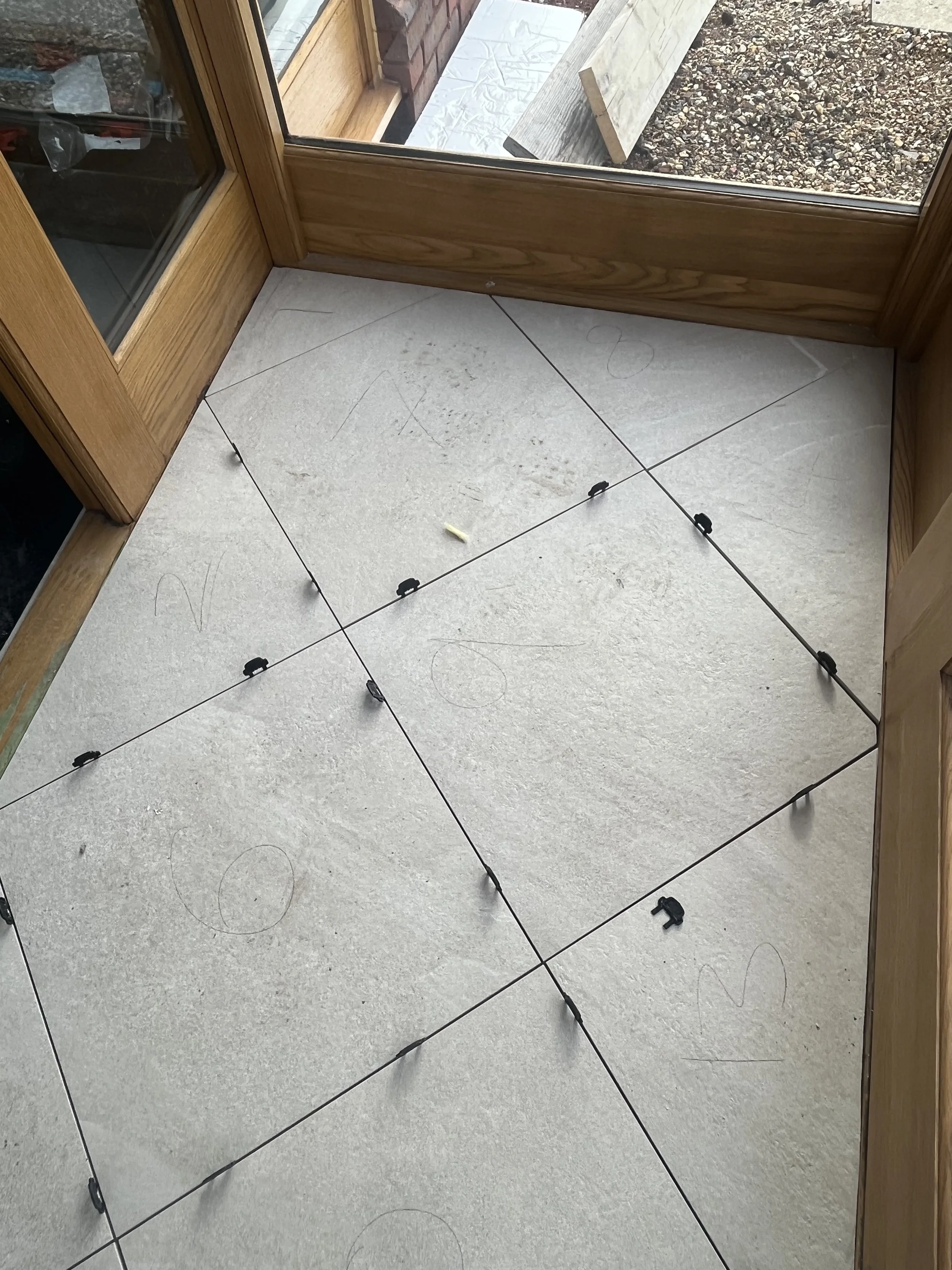 A floor with large square tiles being prepared for installation, with spacers inserted at tile joints, and measurements or markings in pencil on the tiles.