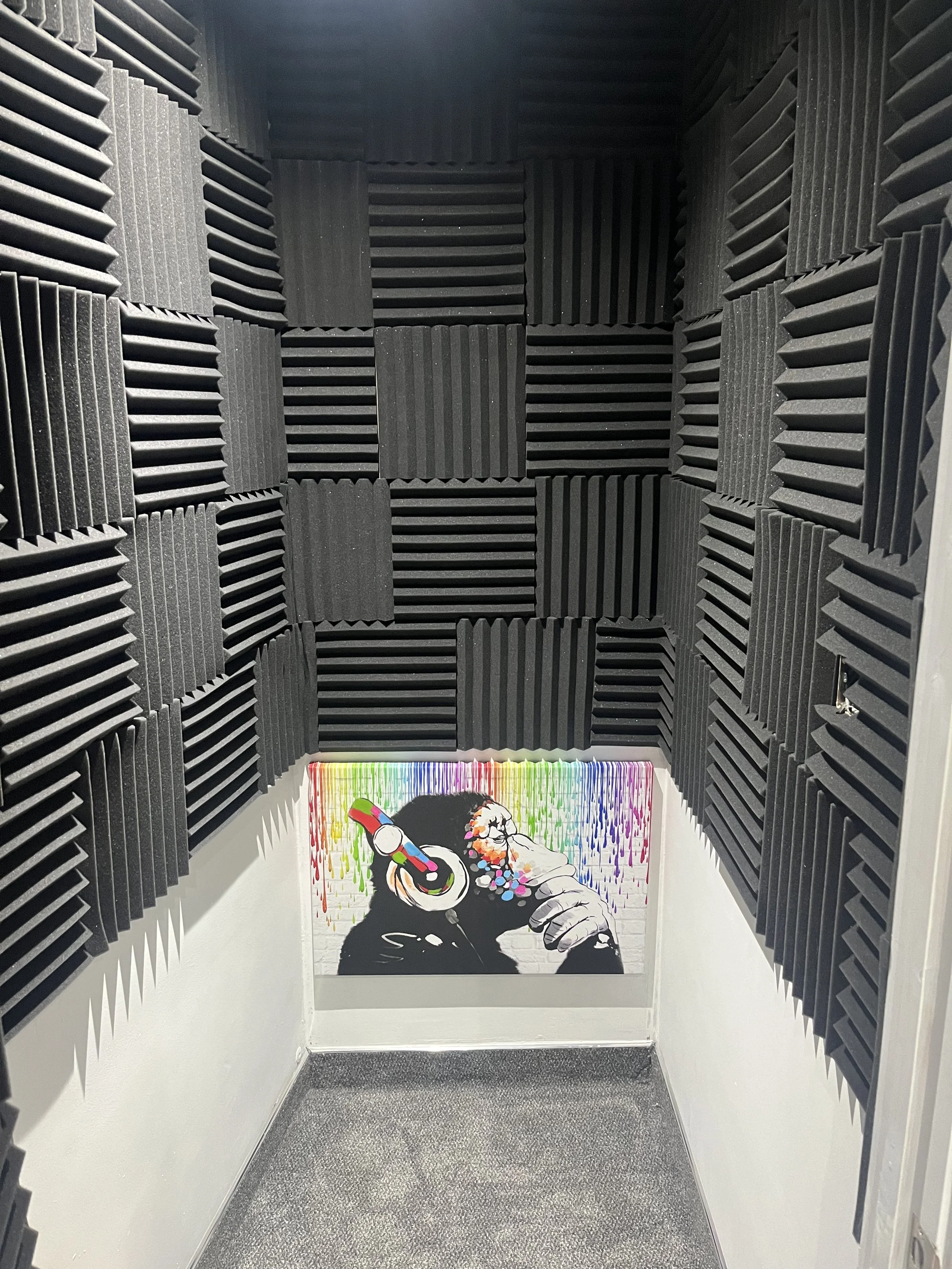 Small soundproof booth with black acoustic foam panels on the walls and colorful graffiti art of a person wearing headphones on the far end wall.