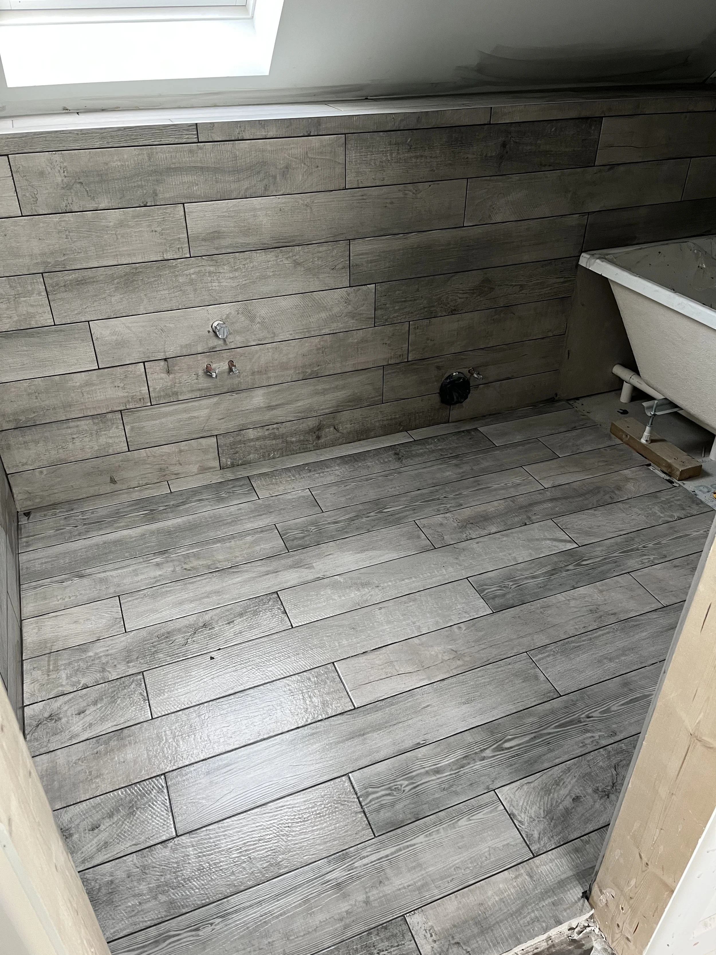 A bathroom under renovation with newly installed grey wood-like floor and wall tiles, a bathtub on the right, exposed plumbing, and an opening in the wall for fixtures.