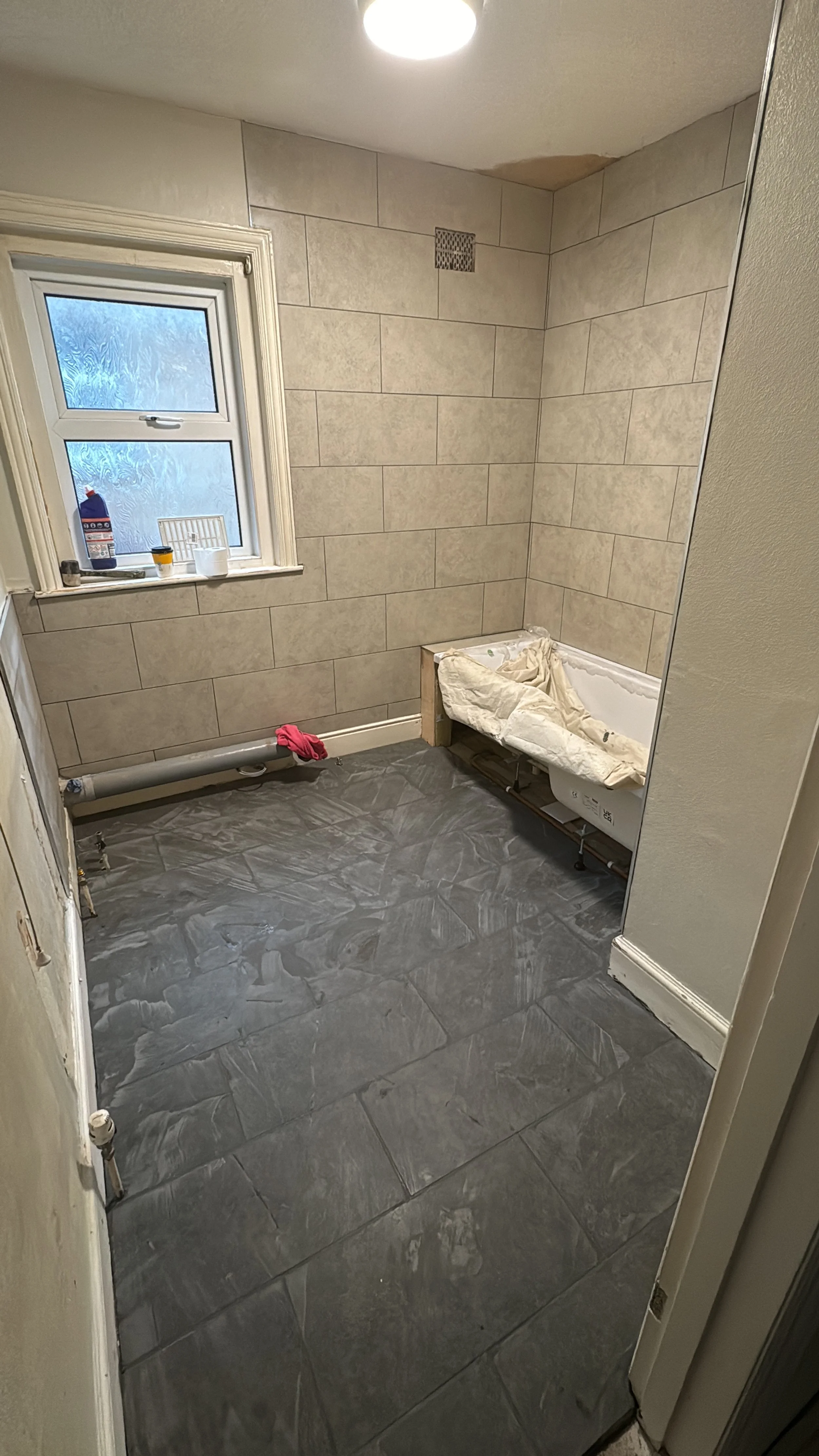 A bathroom under renovation with beige tiled walls, a window with frosted glass, a gray tiled floor, and a bathtub being installed. Construction materials and tools are visible.