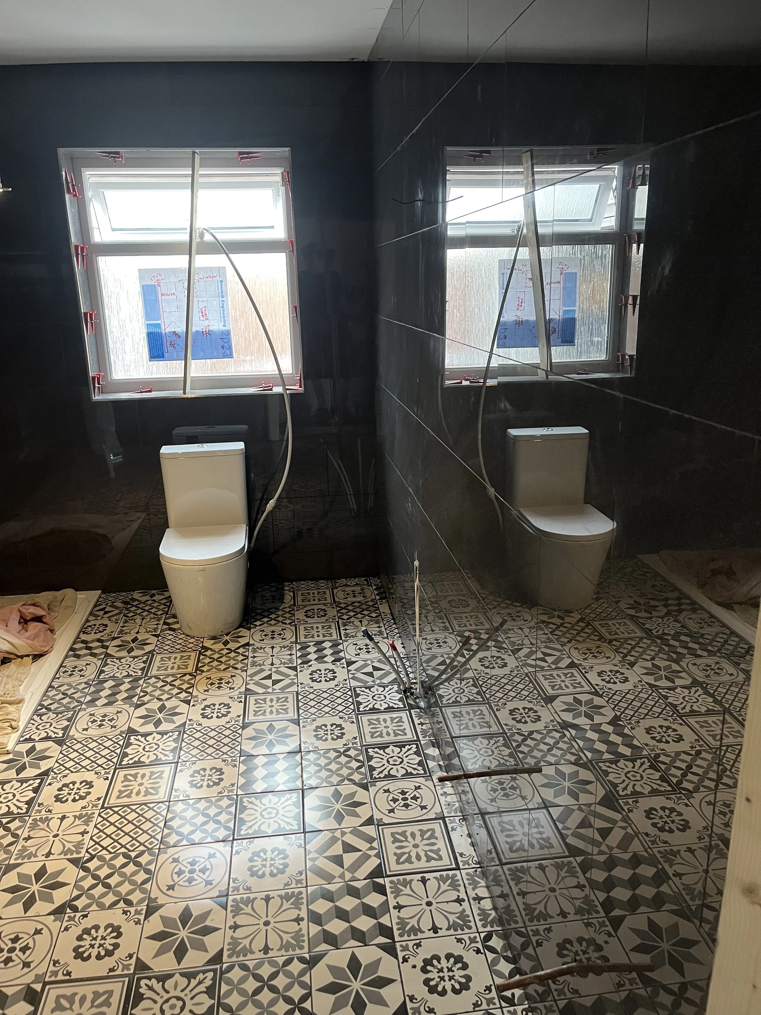 Bathroom under construction with black tiled walls, patterned tile floor, two windows, a ceiling-mounted extendable shower device, and two modern toilets, one on each side of the room.