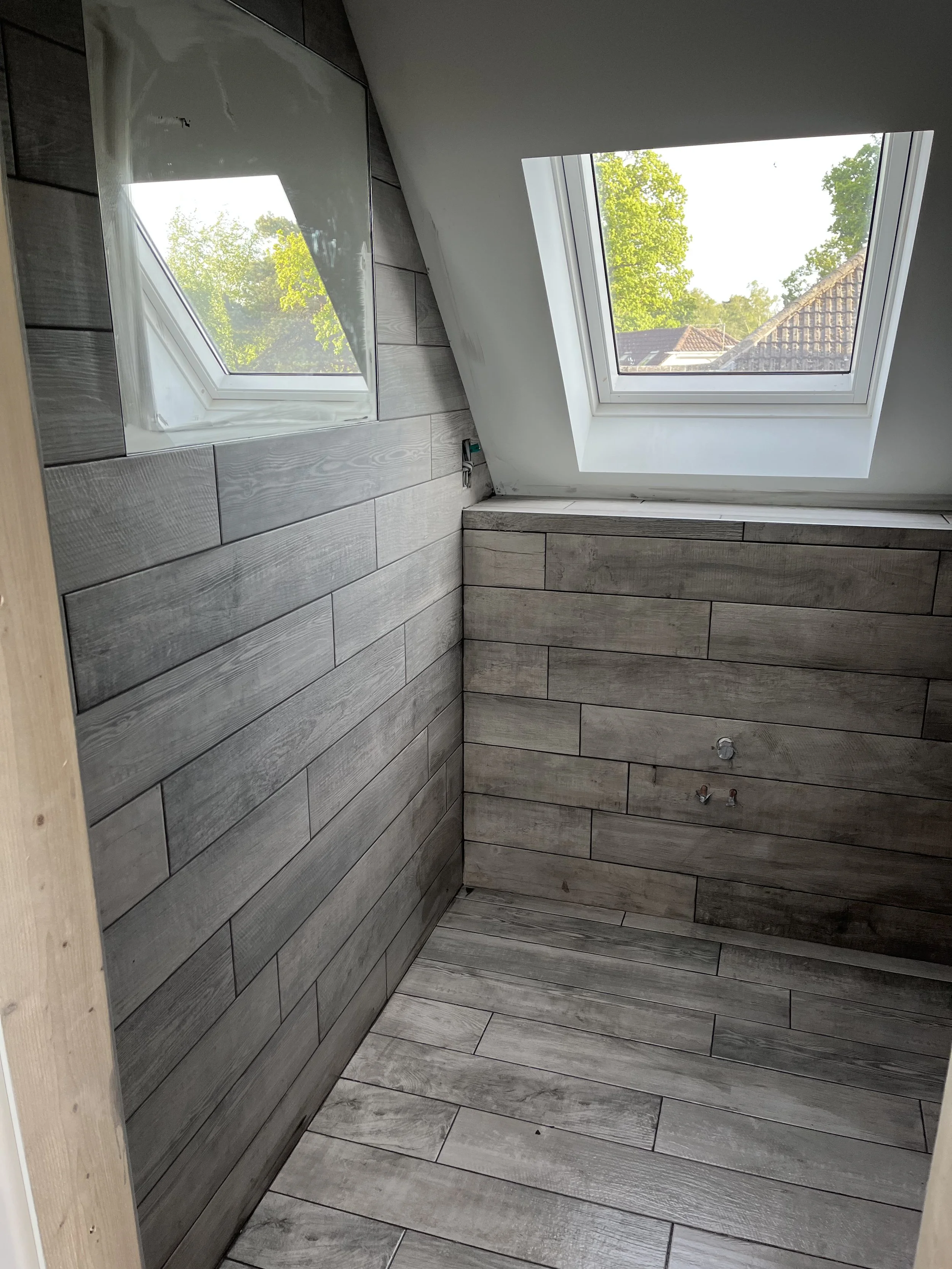 An unfinished bathroom with wooden wall and floor tiles, windows on slanted ceiling showing trees and rooftops outside, plumbing visible on the wall.