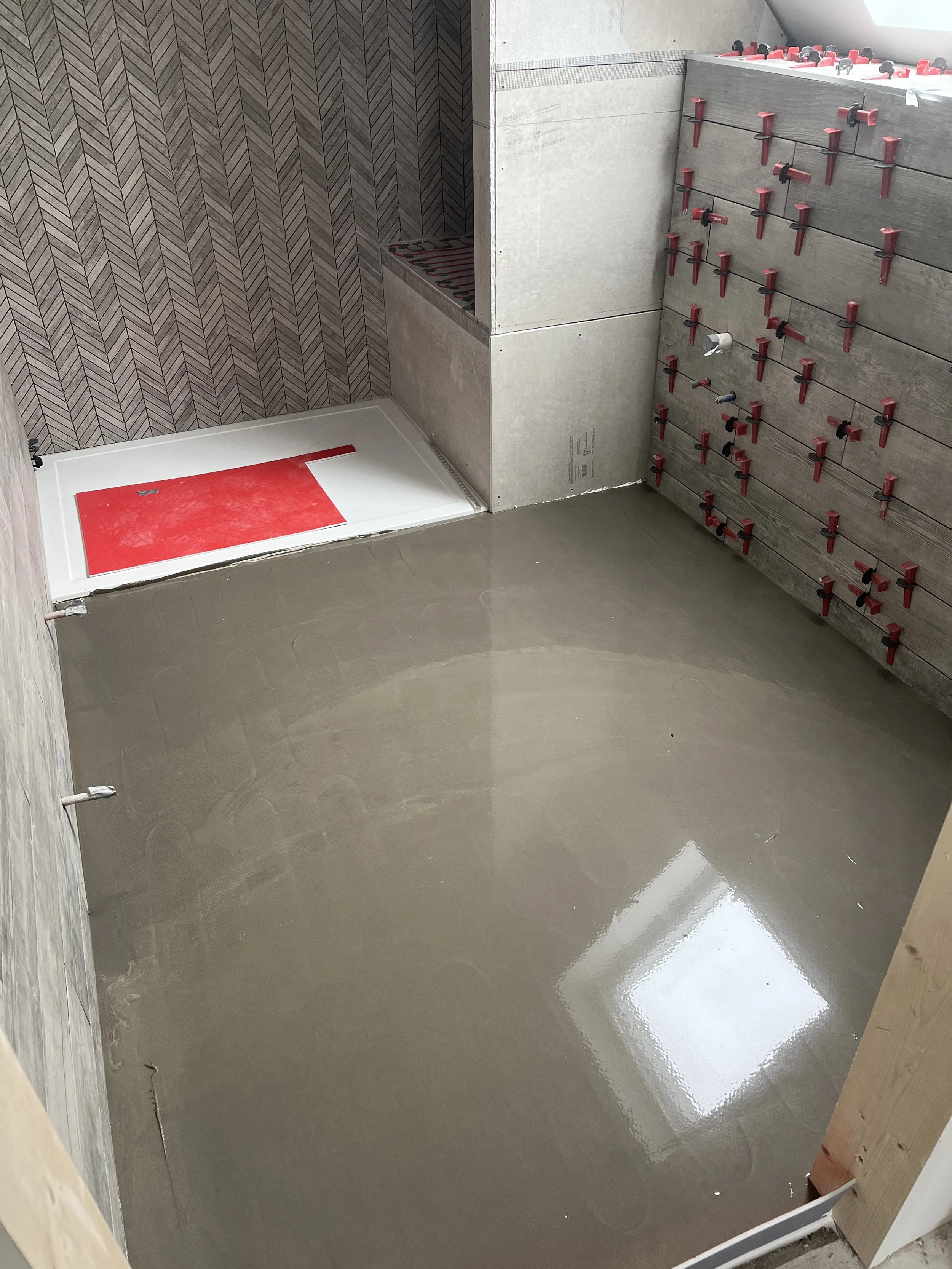 Bathroom under construction with freshly poured concrete flooring, a shower area with red-backed wall tiles, and a wall with multiple red and black plumbing fittings.