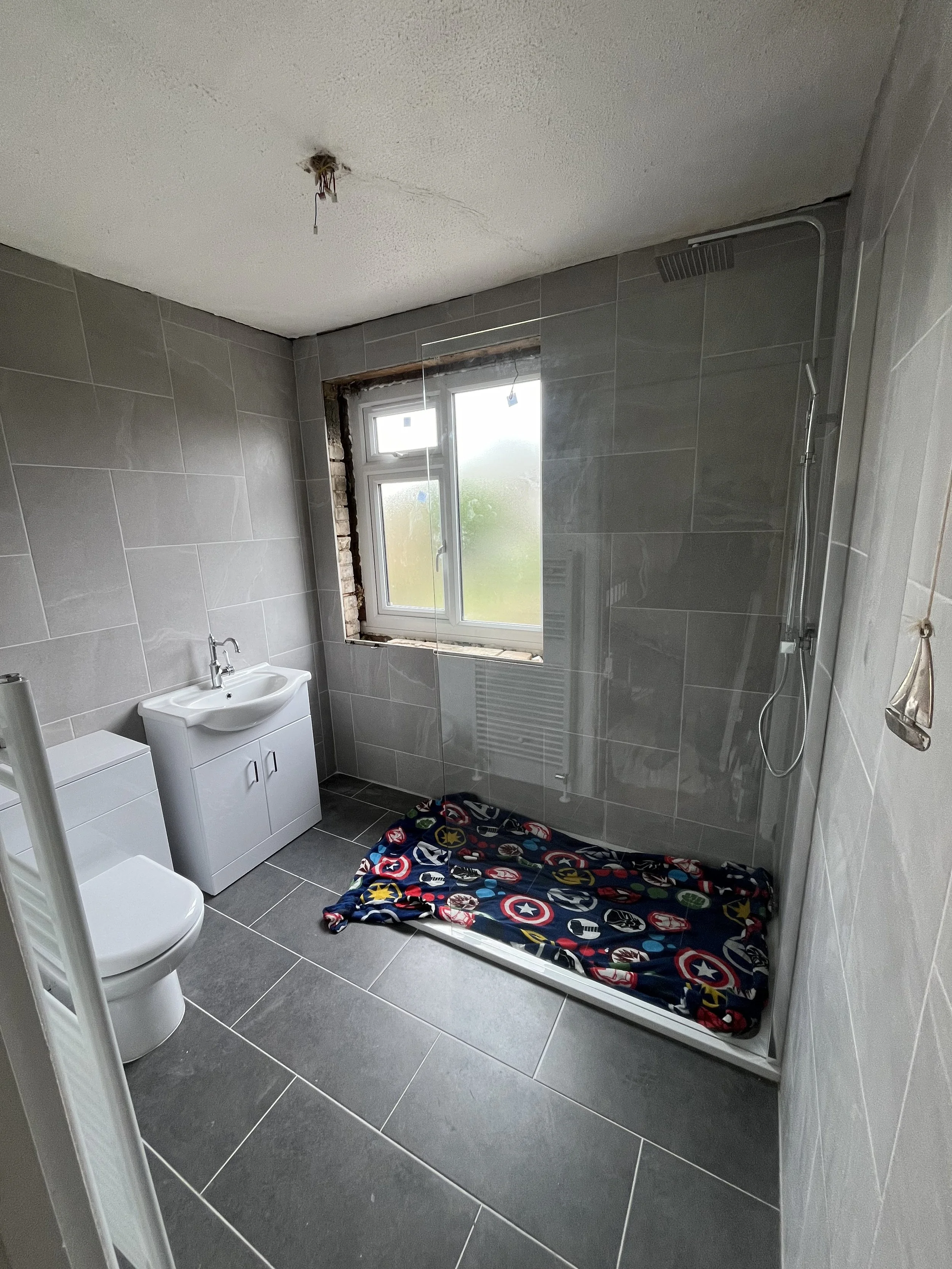 A partially finished bathroom with gray tiled walls and floor, a white toilet, a small white sink cabinet, a large frosted window, and a shower area with a black superhero-themed blanket on the shower tray.