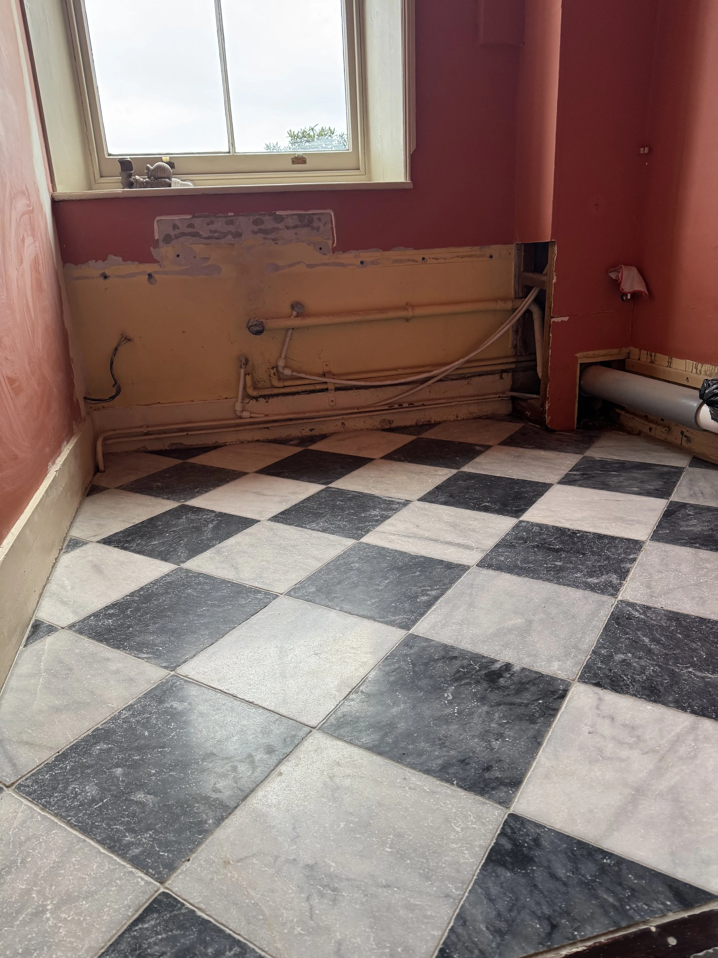 A room under renovation with black and white checkered tile flooring, a window, and exposed plumbing on the wall.