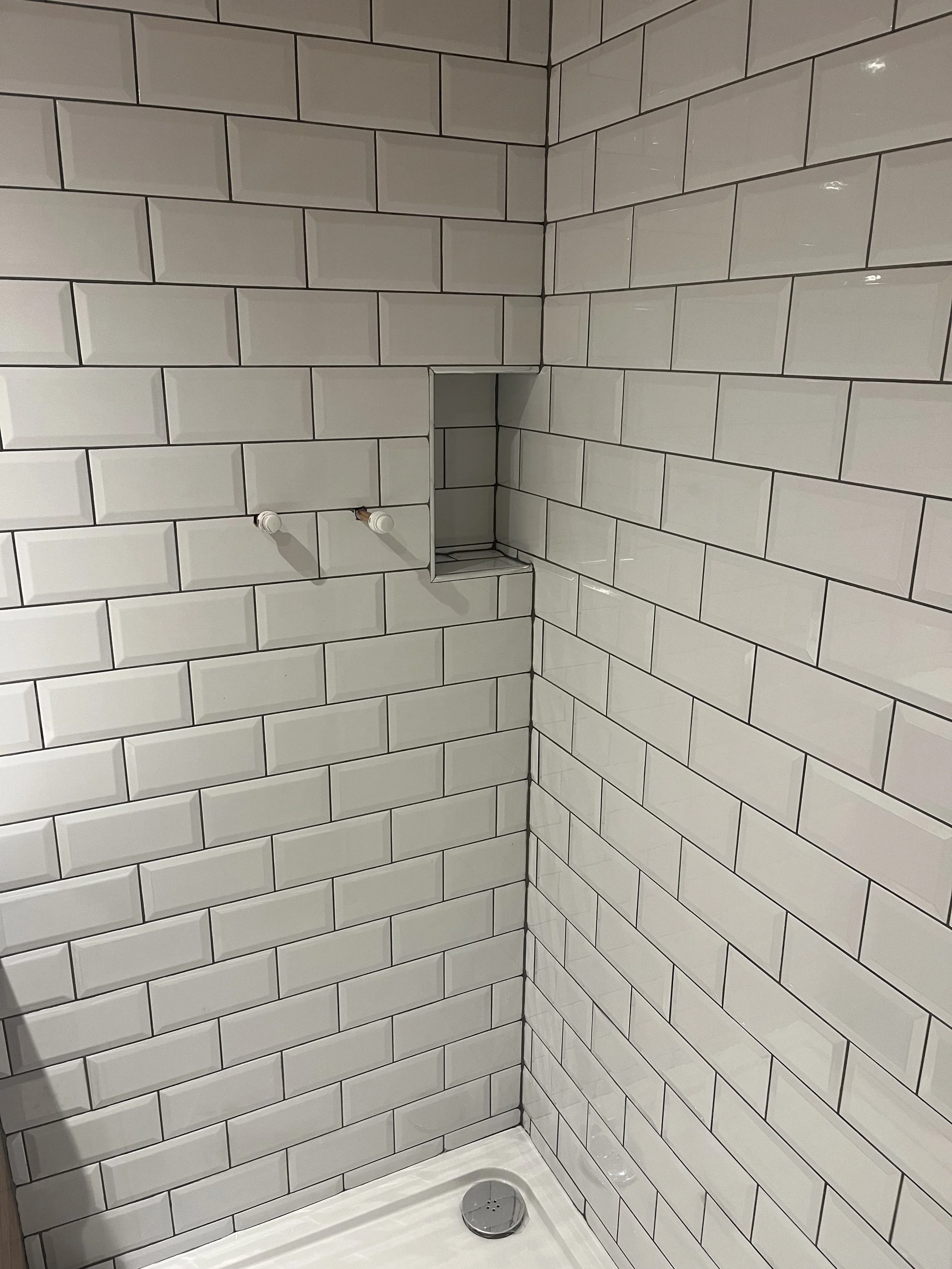 Empty shower with white subway tiles, a niche in the wall, two protruding faucet connections, and a round drain on the floor.