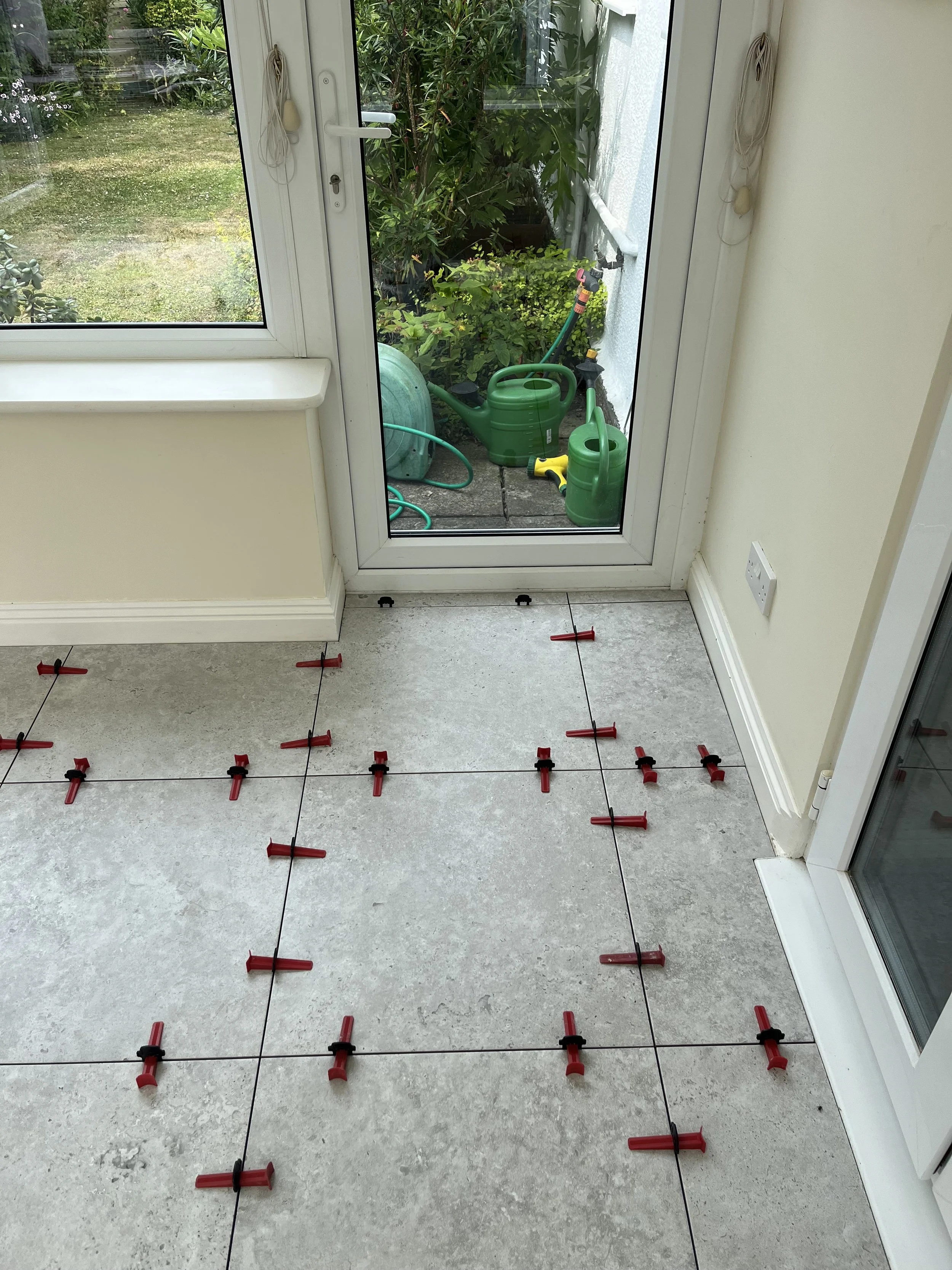 Tile floor being measured and marked with red tile spacers for installation, near a glass door leading outside to a garden area with watering cans and gardening tools.