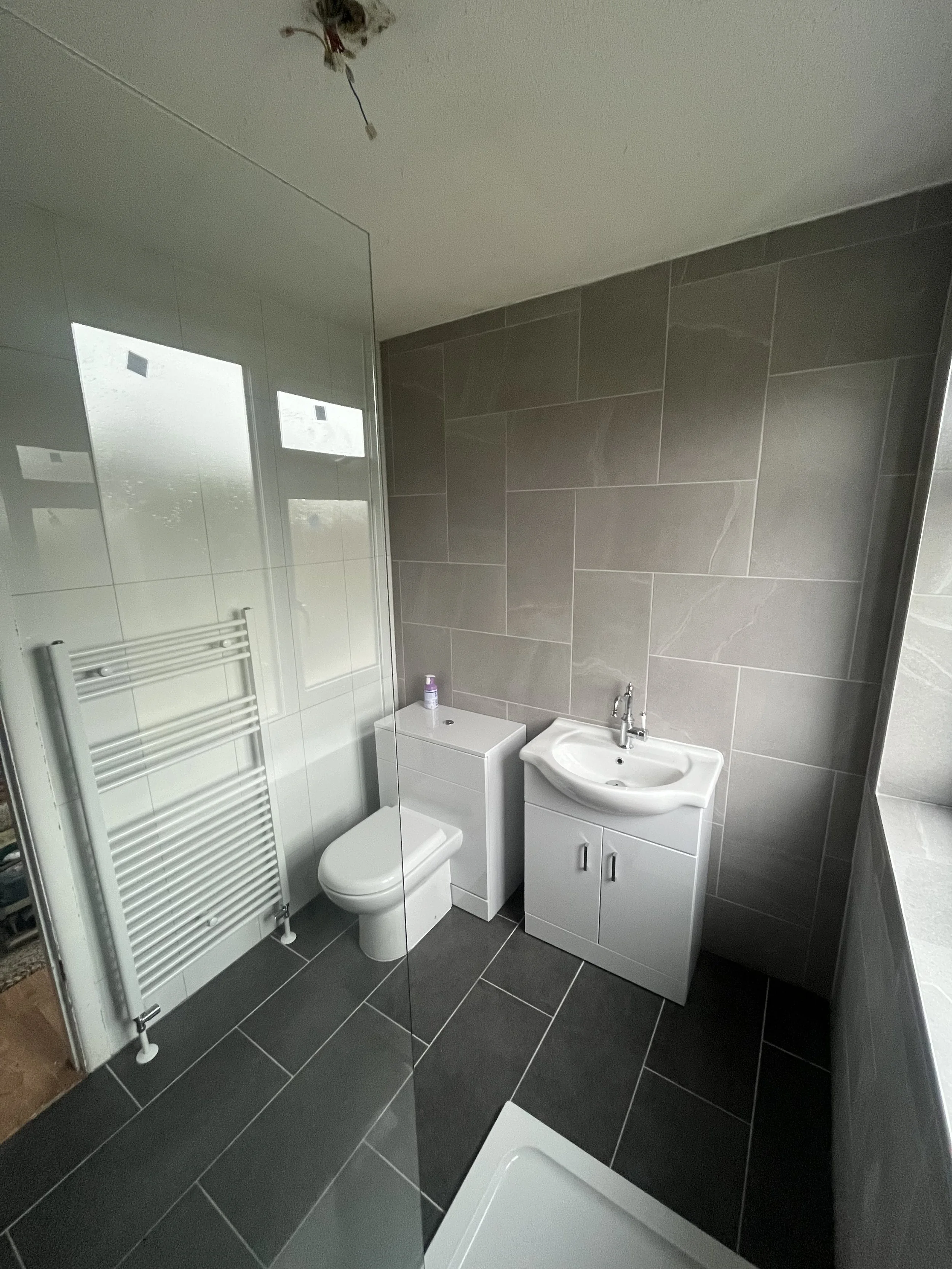 A modern bathroom with gray tiled walls and floors, a toilet next to a small white cabinet, a sink with a cabinet below, and a heated towel rail on the wall, partially visible shower area, and a large window letting in natural light.