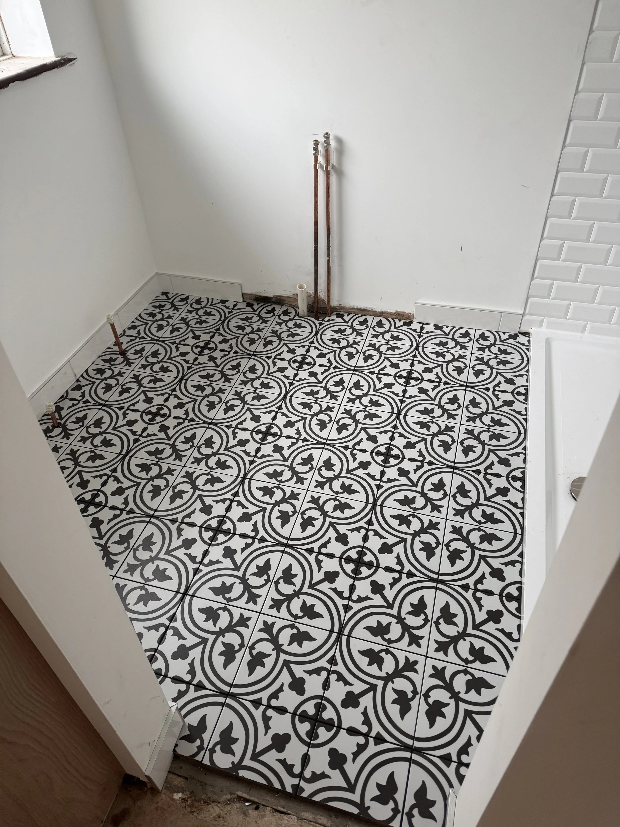 A small room with decorative black and white patterned floor tiles, with visible plumbing pipes along the back wall, and a partially finished white wall with a section of subway tile on the right.