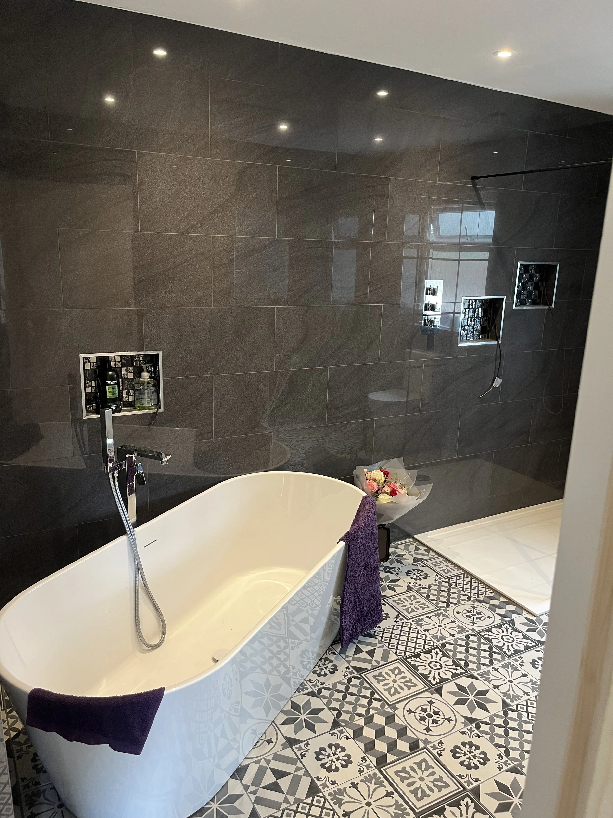 Modern bathroom with a white freestanding bathtub, purple towels hanging on it, a bouquet of flowers on the floor, dark tiled wall, patterned tile floor, and built-in shower niches.