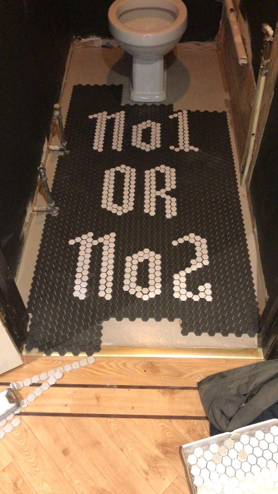 A tiled bathroom floor with black and white tiles forming the words 'NO OR 10.2' leading to a toilet. The side walls are partly unfinished, and a small white tiled mat is visible at the bottom right corner.