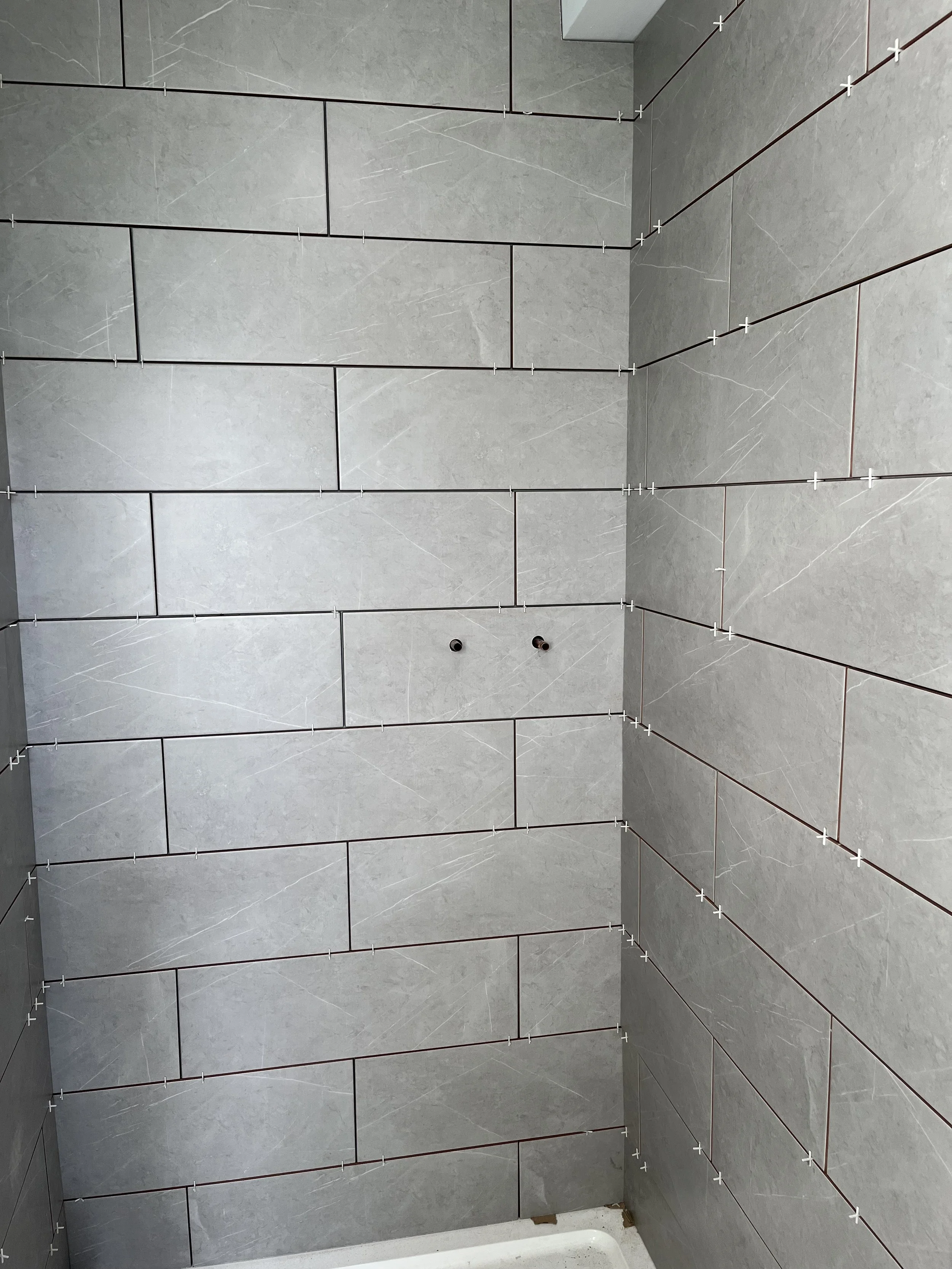 Bathroom wall with gray rectangular tiles installed and tile spacers in place, with two plumbing holes visible.