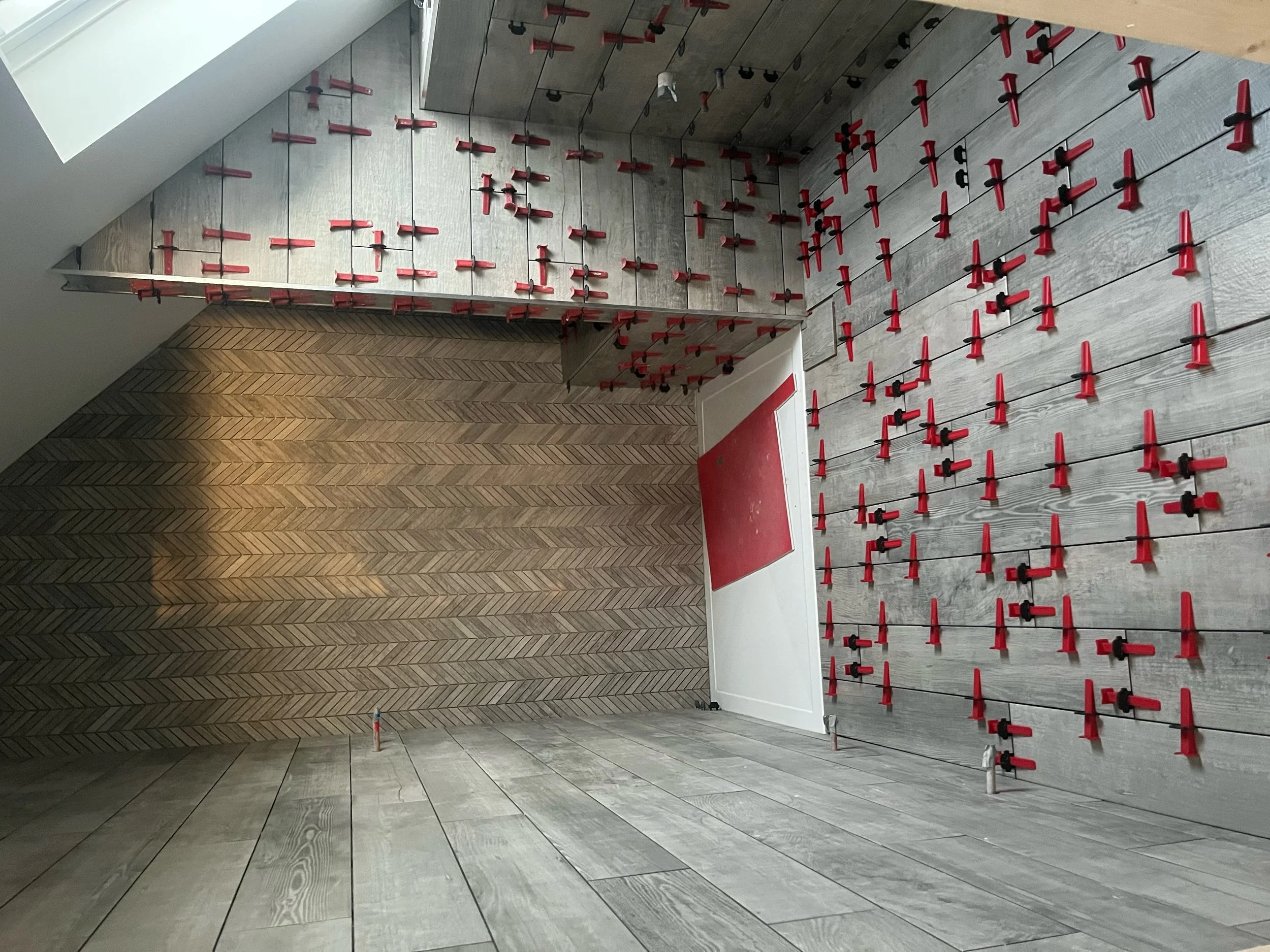 Interior of a room under construction with walls covered in horizontal wood panels and numerous red clamps fastening the wall panels; the floor has light-colored wood planks and a red carpet near the door.