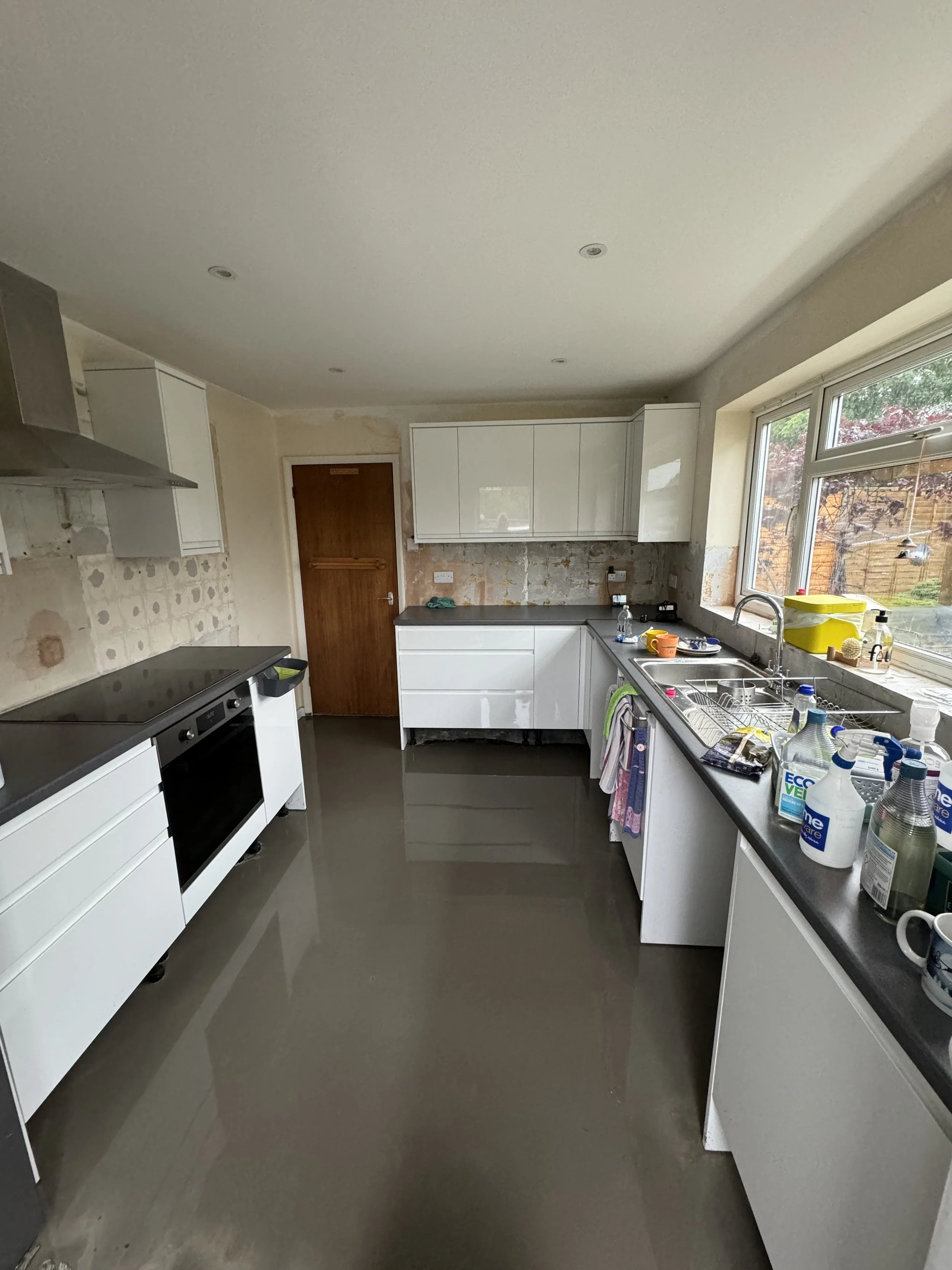 Kitchen with white cabinets, black countertops, and a large window; appears to be under renovation or cleaning, with cleaning supplies and dishes on the counter.
