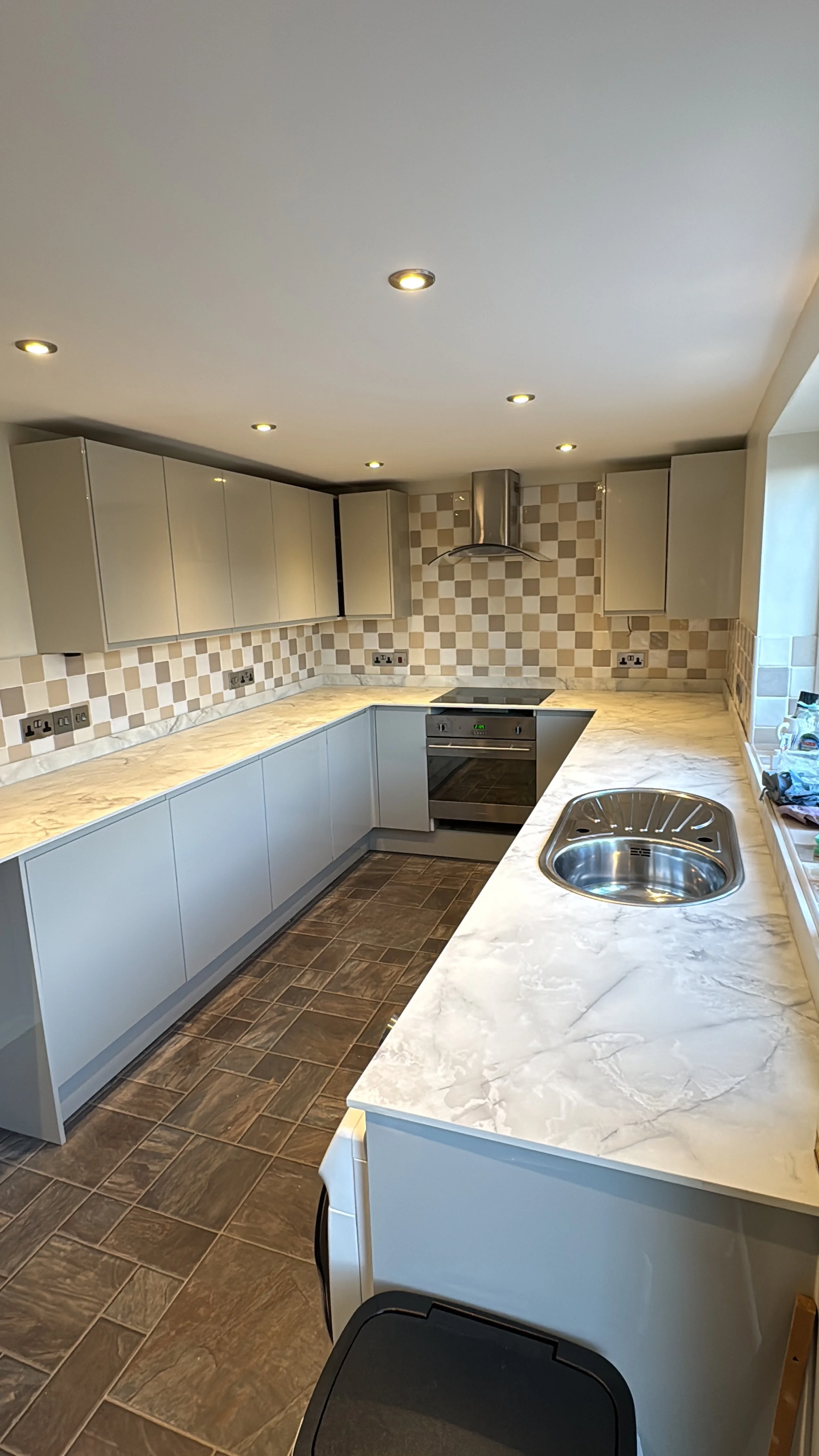 Empty modern kitchen with marble countertops, beige cabinets, tiled backsplash, stainless steel oven and range hood, and a round sink.
