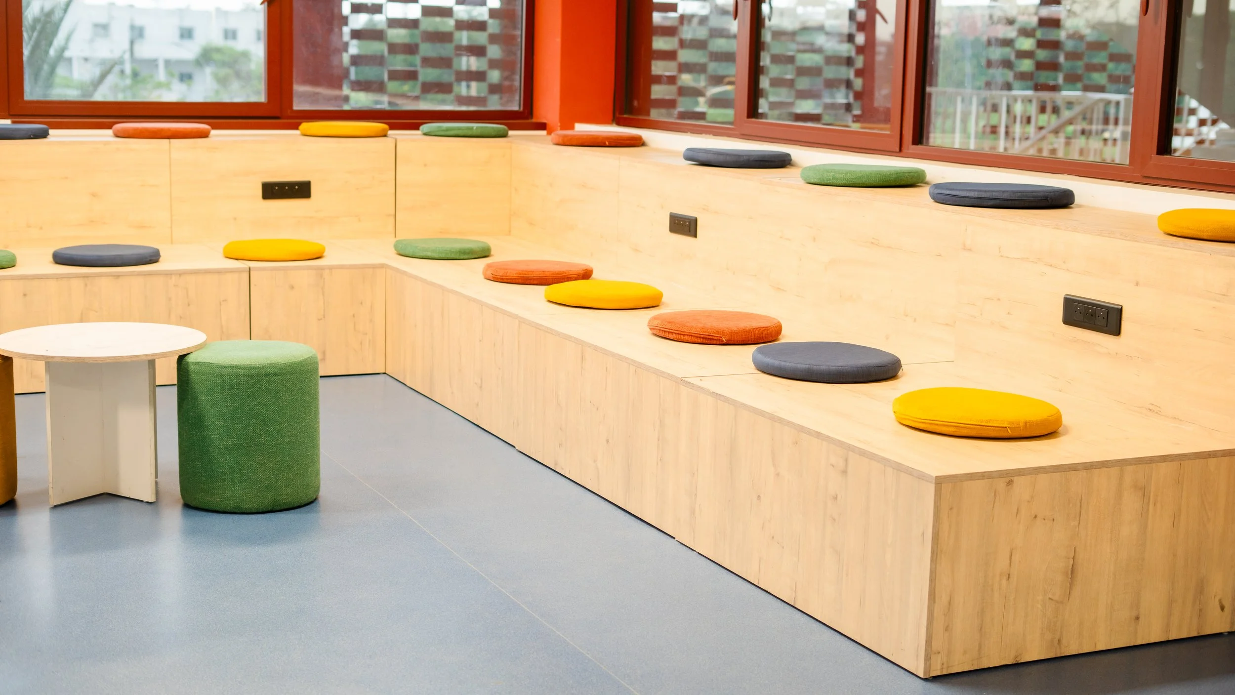 espace de détente avec un banc en bois clair, coussins de différentes couleurs, et un petit tabouret vert et une petite table blanche. sheznou
