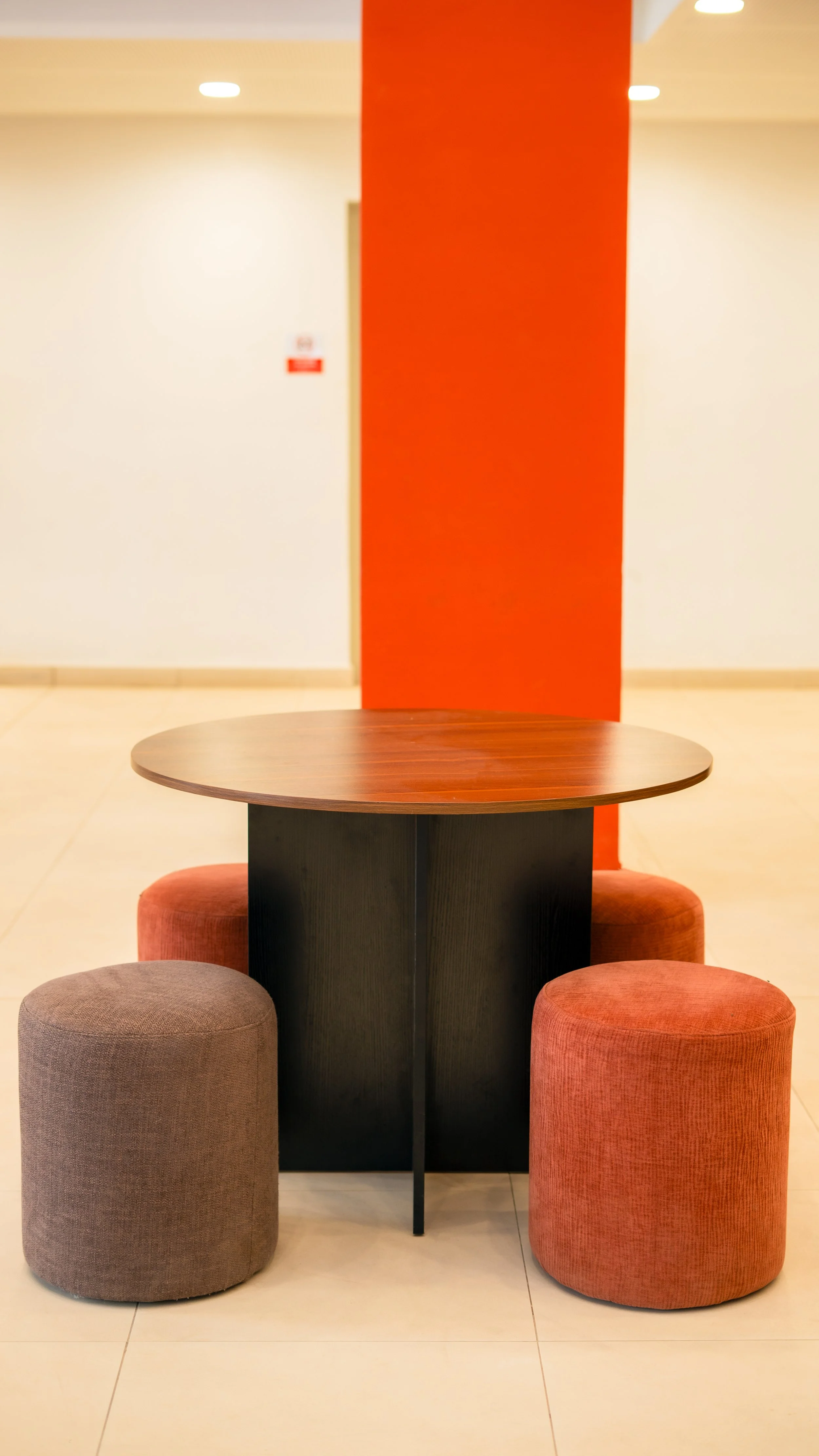 Table ronde en bois entourée de deux ottomans en tissu, un gris et un rouge, en face d’un mur avec une colonne rouge au centre. sheznou