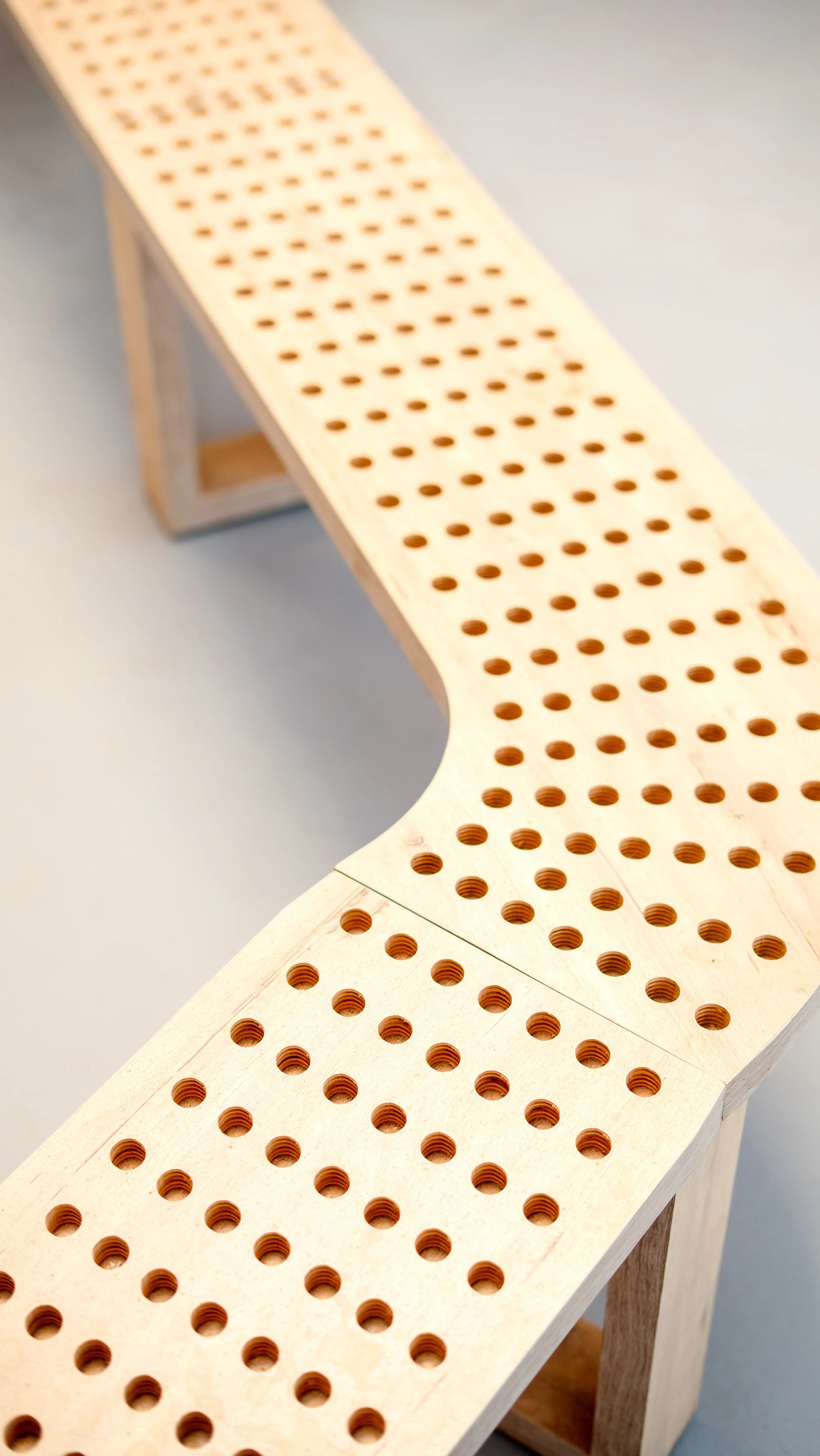 Banquette en bois clair avec plusieurs trous ronds dans le dossier et l'assise, disposée sur un sol gris. sheznou