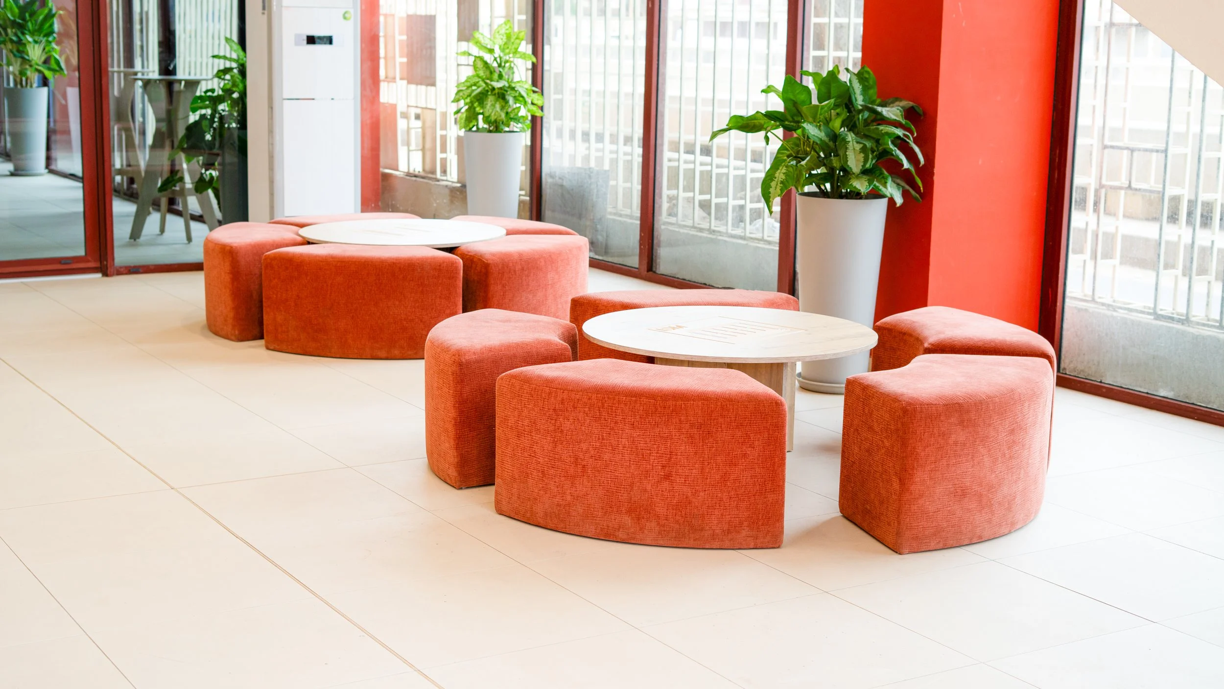 Espace de détente avec des fauteuils orange et des tables rondes, près de grandes fenêtres avec des plantes en pot. sheznou