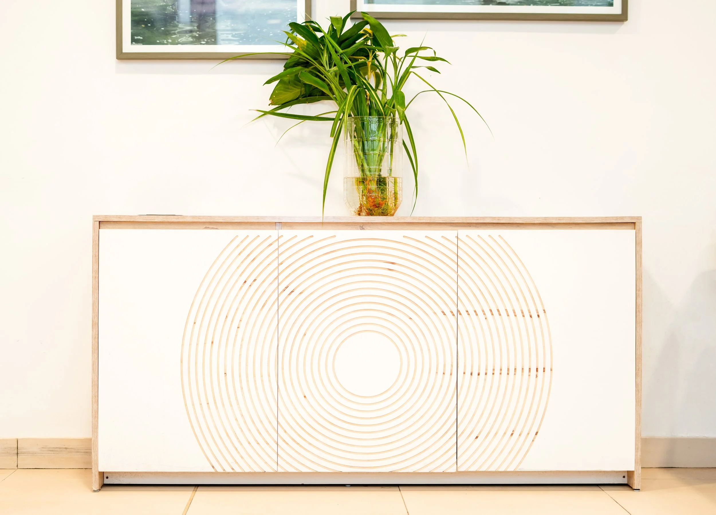Vase avec des plantes vertes sur une commode blanche en bois avec un motif circulaire. Photo encadrée au mur. sheznou