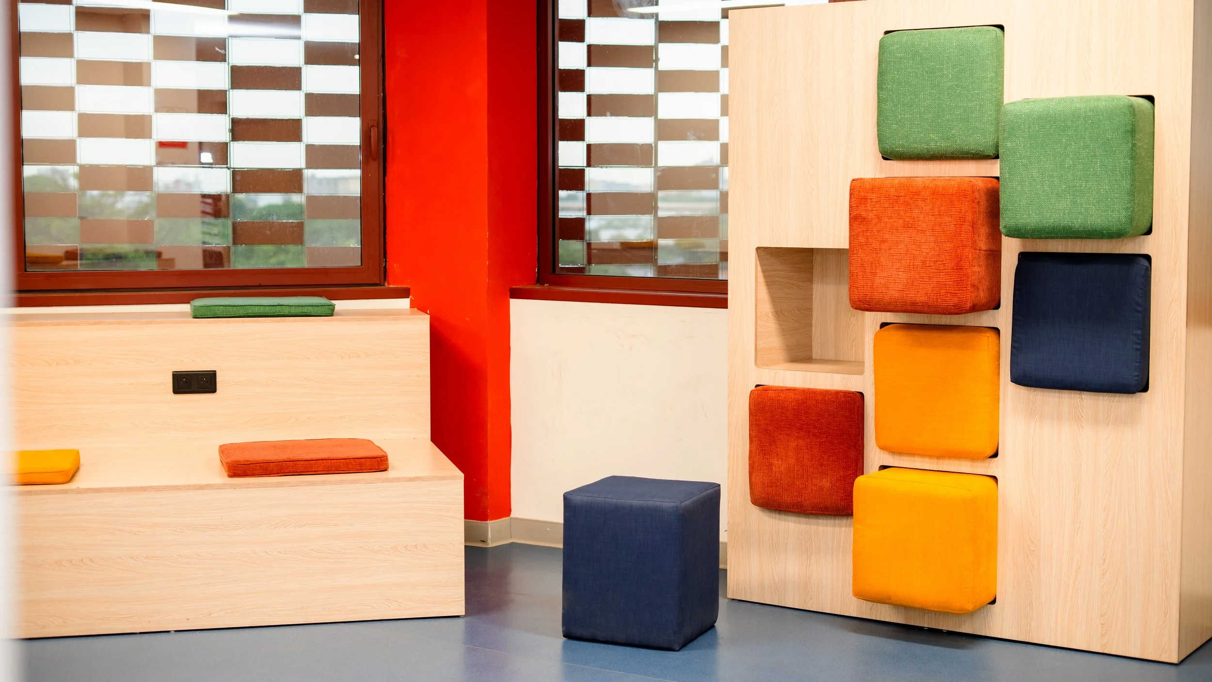 Intérieur avec des sièges colorés en bois clair, comprenant des coussins verts, oranges, jaunes et bleus, devant une grande fenêtre avec volets à la française. sheznou