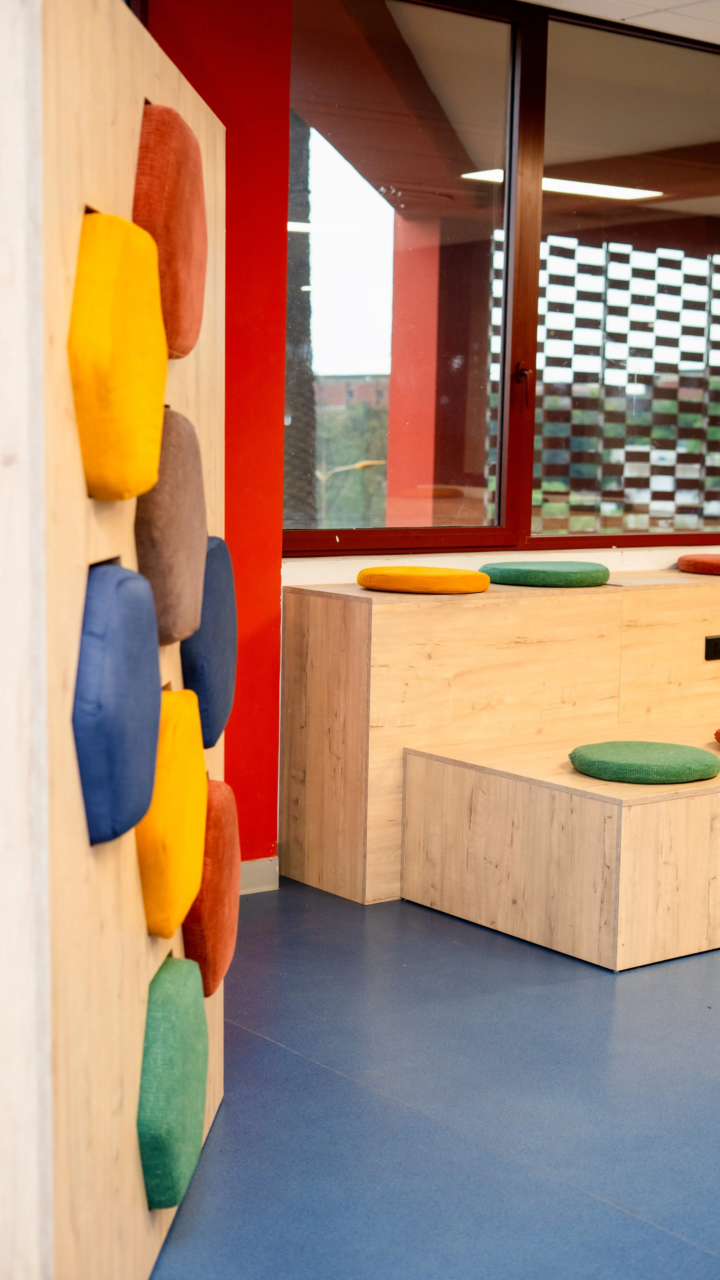 Intérieur coloré avec des coussins en tissu vert, jaune, rouge, beige et bleu, sur un meuble en bois clair et un mur en bois avec des coussins en forme de cailloux. sheznou