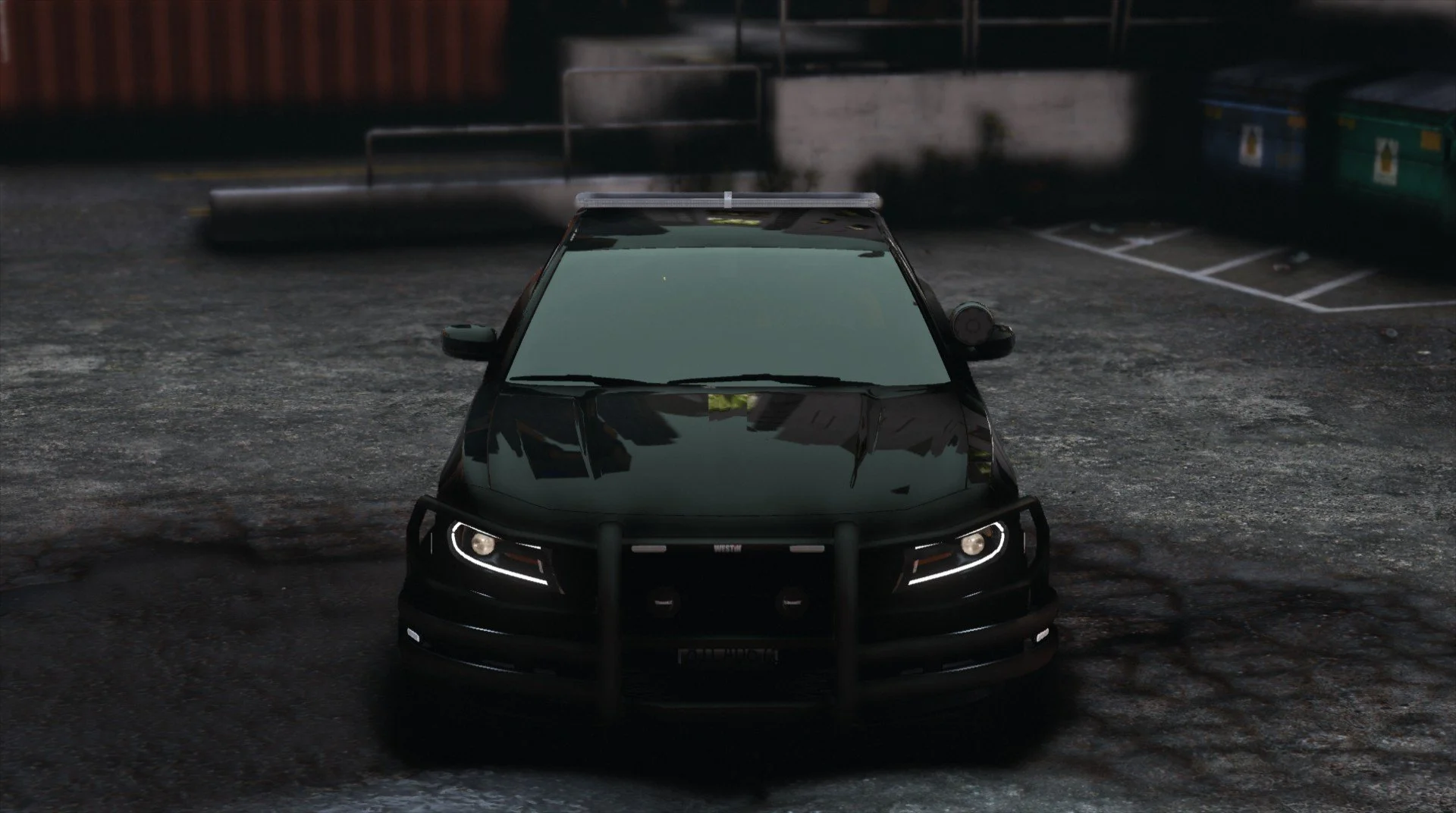 Black police car with a push bumper, parked on a gravel lot at night, with containers and a building in the background.