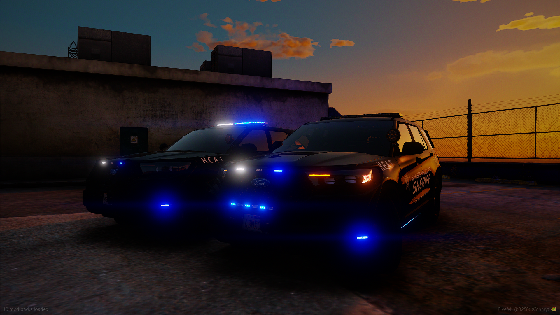 Two police cars with flashing lights parked on a rooftop parking lot at sunset, with a fence and buildings in the background.