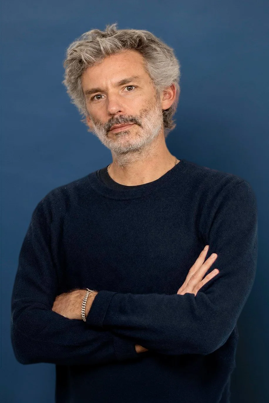 Portrait d'un homme d'âge moyen avec des cheveux gris, une barbe et une expression neutre, portant un pull noir avec un fond bleu.