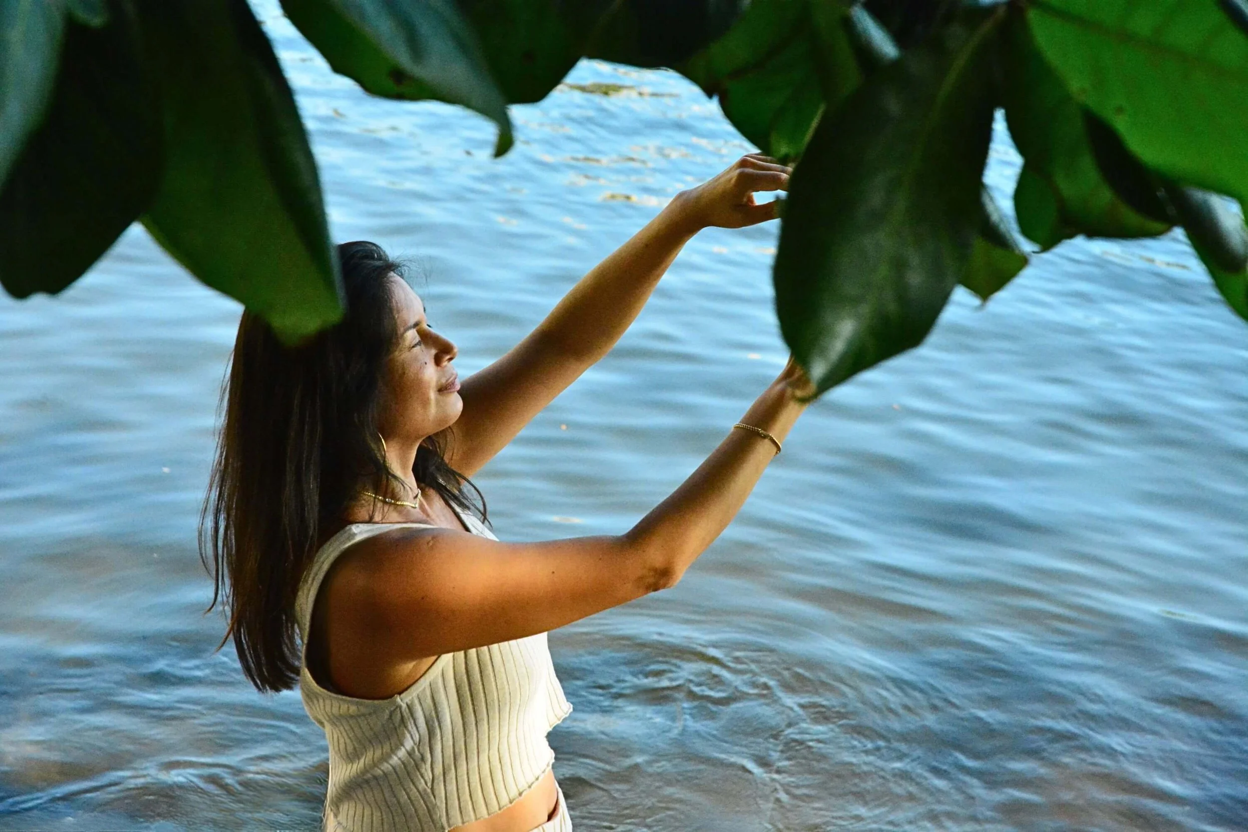 A woman is standing in the water by a river or lake, raising her right hand to reach for leaves on a tree or shrub, while the water ripples softly in the background.