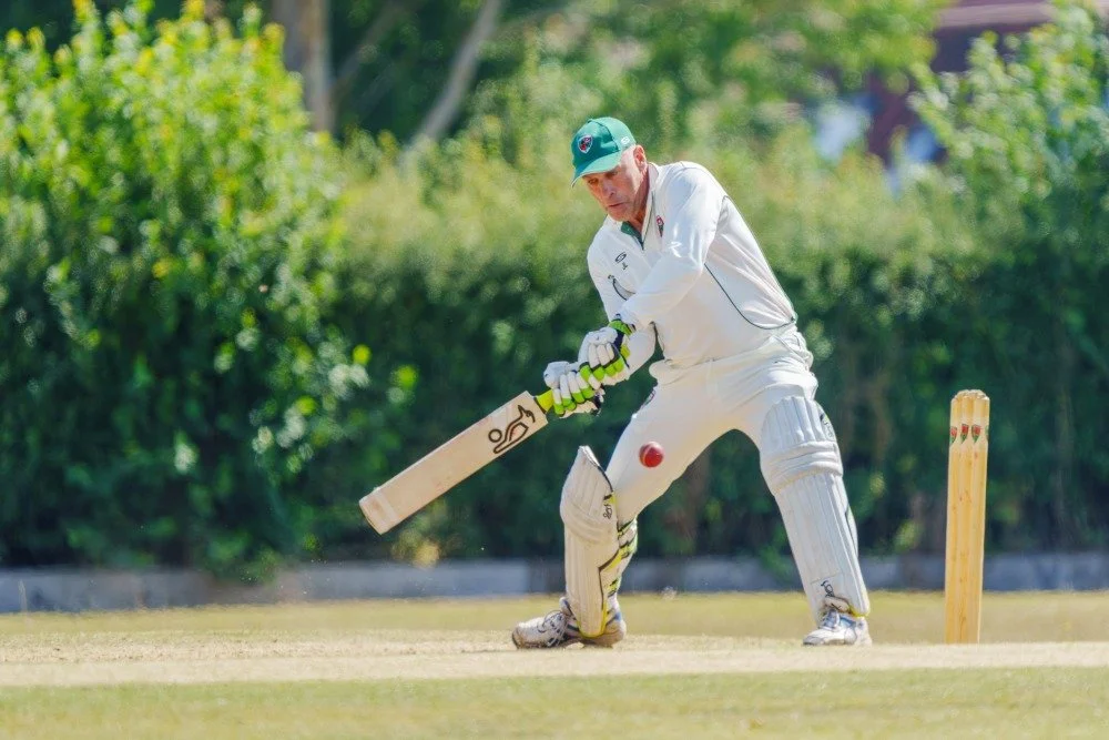 A cricket player in white uniform and helmet ready to hit a ball with a cricket bat on a field with green trees in the background.