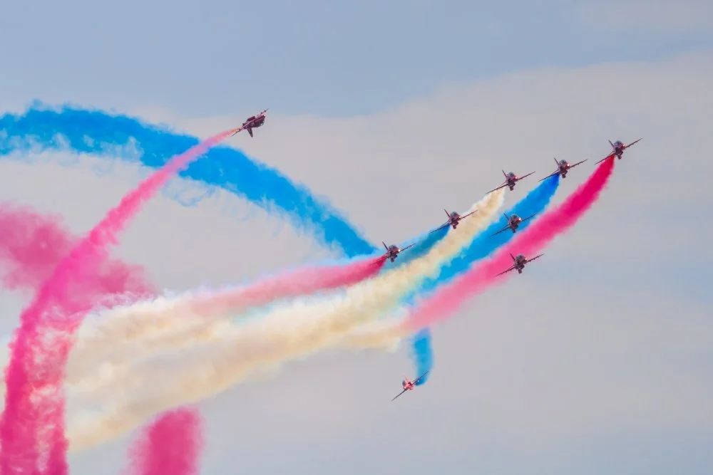 Multiple airplanes performing an aerial stunt, releasing colorful smoke trails in blue, pink, and yellow across the sky.