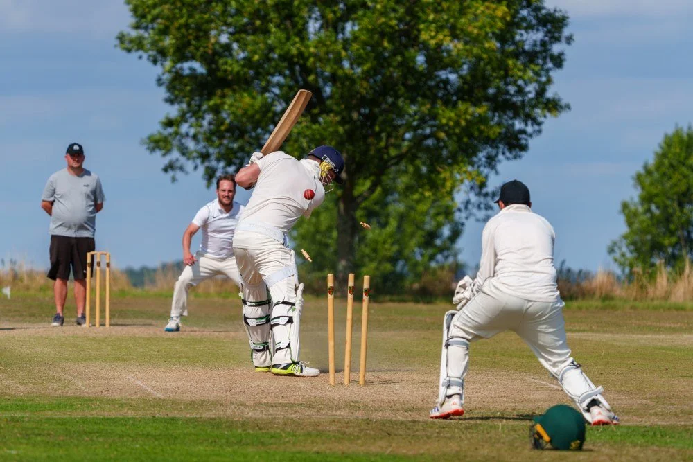 Cricket players on the pitch with a batsman swinging the bat, a wicketkeeper preparing to catch, and two other players watching in the background outdoors.