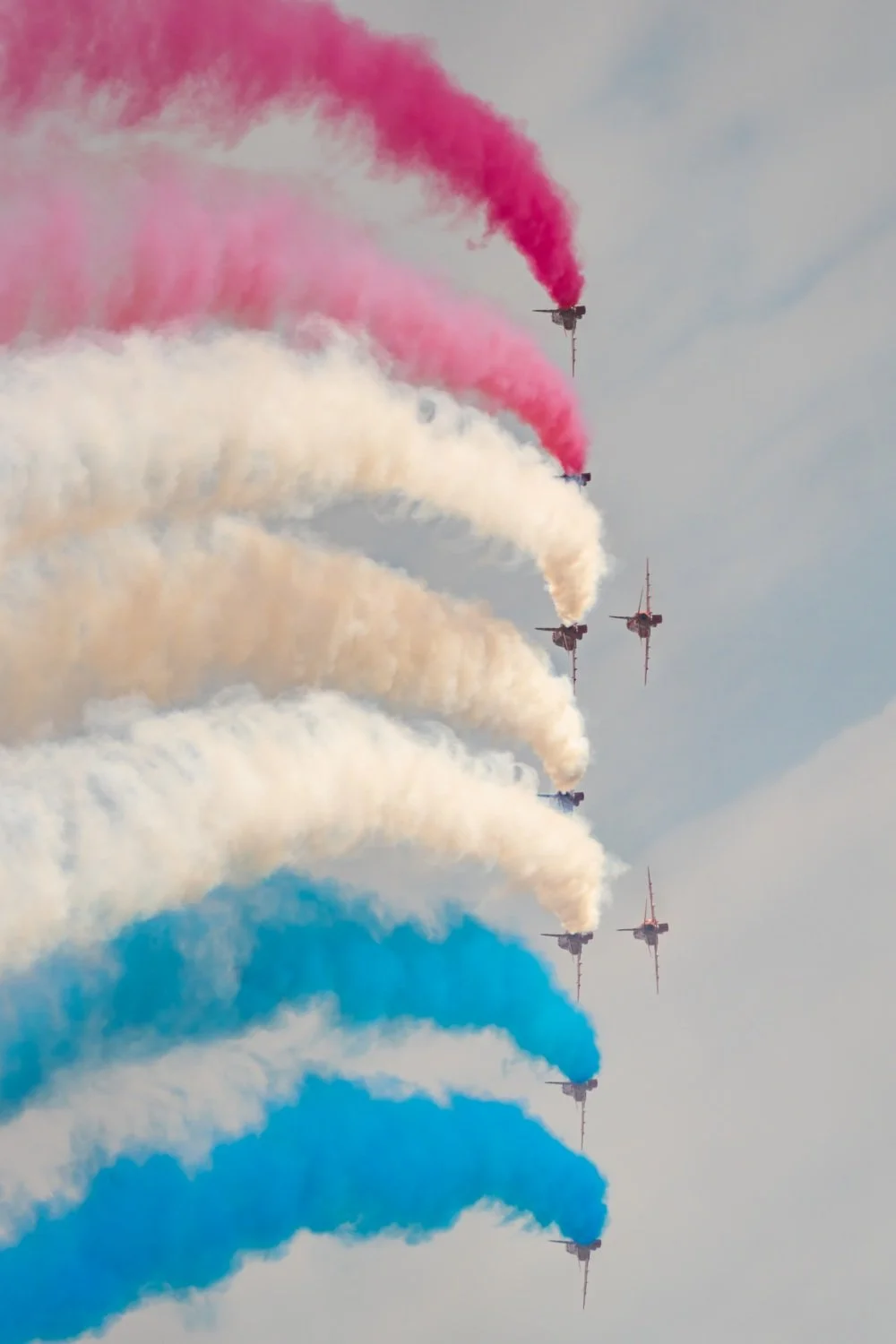 Five airplanes flying in formation releasing colored smoke trails in pink, white, and blue against a cloudy sky.