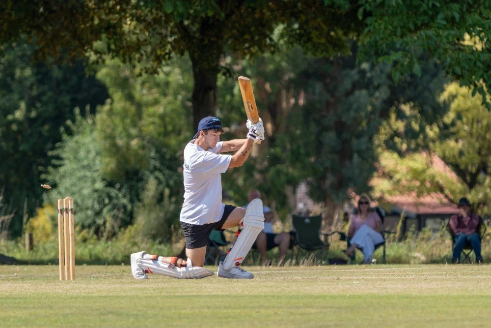 A man batting in a cricket match outdoors on a grassy field with trees and spectators in the background.