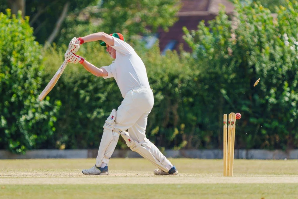 A person in white cricket attire batting on a cricket field with greenery in the background.