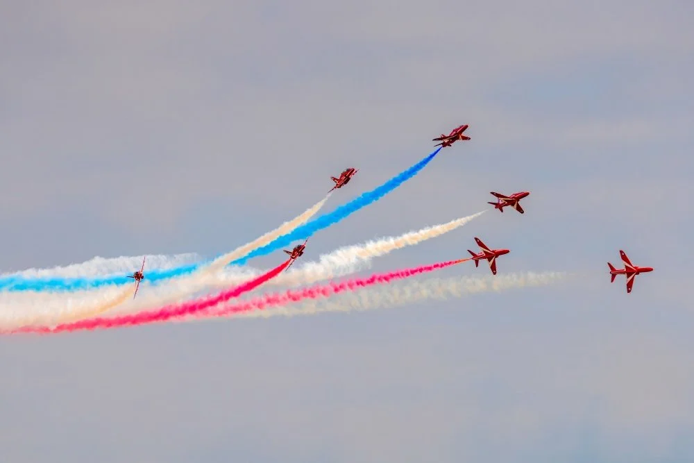 Six red airplanes flying in a formation with red, white, blue, and pink smoke trails in the sky.