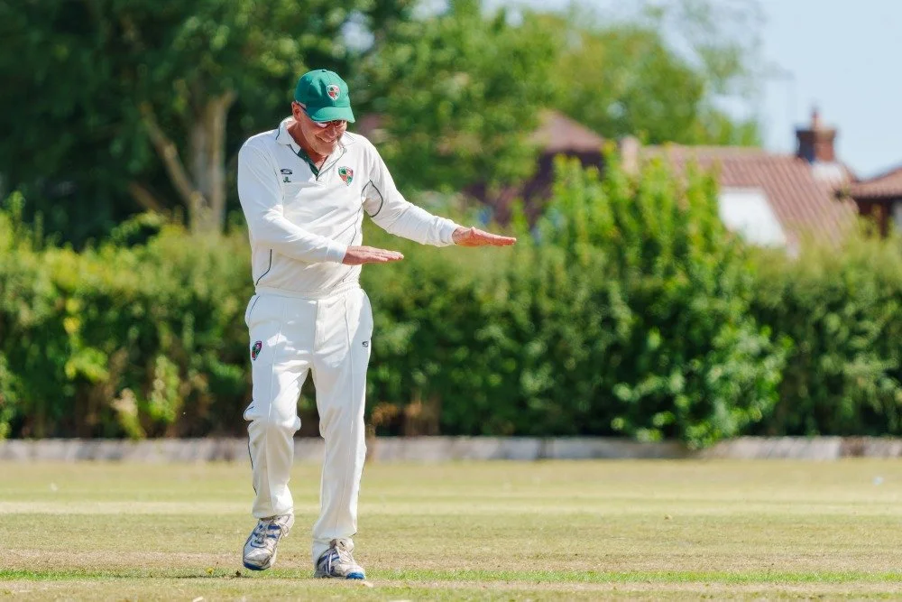 A man dressed in white cricket uniform and cap, standing on a cricket field, gesturing with his hands, with greenery and houses in the background.