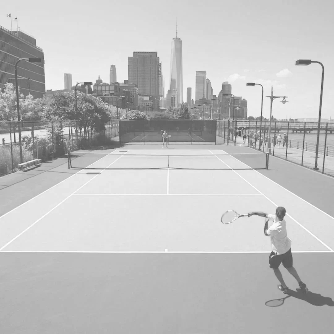 A man playing tennis on an outdoor court near a city with tall skyscrapers, including One World Trade Center, on a sunny day.