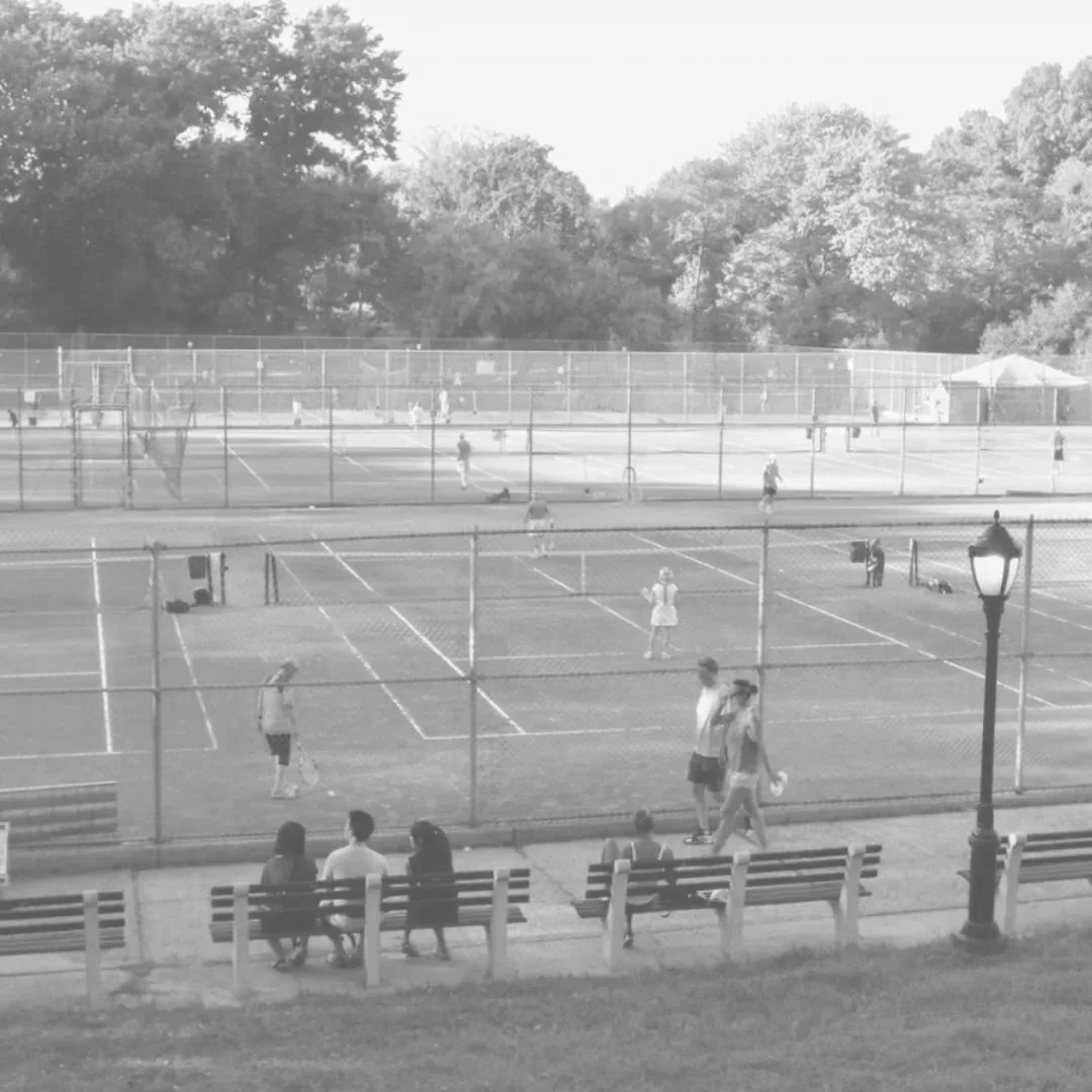 Tennis courts with people playing, sitting on benches, and a lamp post in a park area with trees in the background.