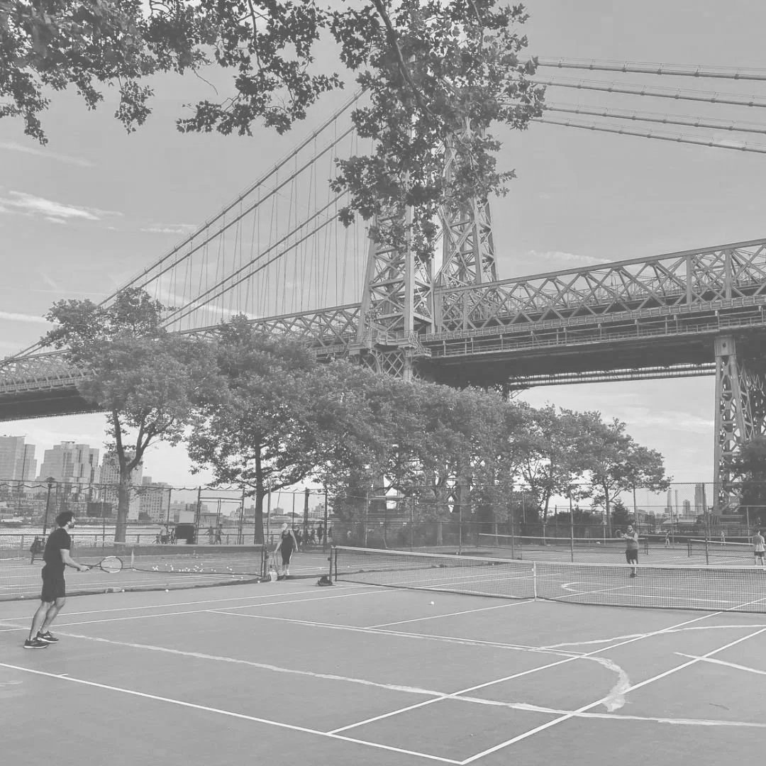 People playing tennis on an outdoor court under a bridge with trees and a city skyline in the background.