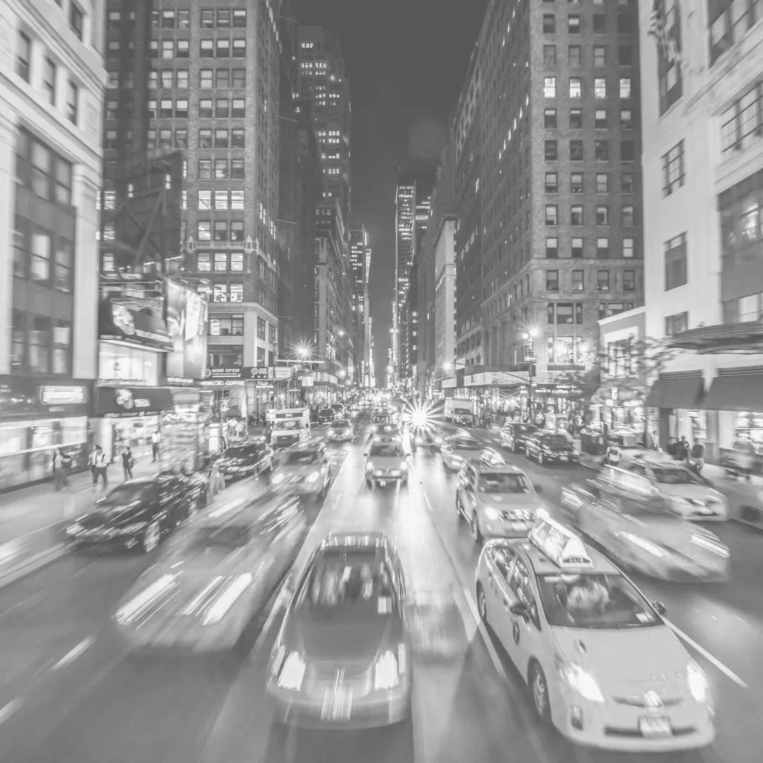 Nighttime city street filled with yellow taxis, cars, and pedestrians, surrounded by tall skyscrapers with bright lights and illuminated storefronts.