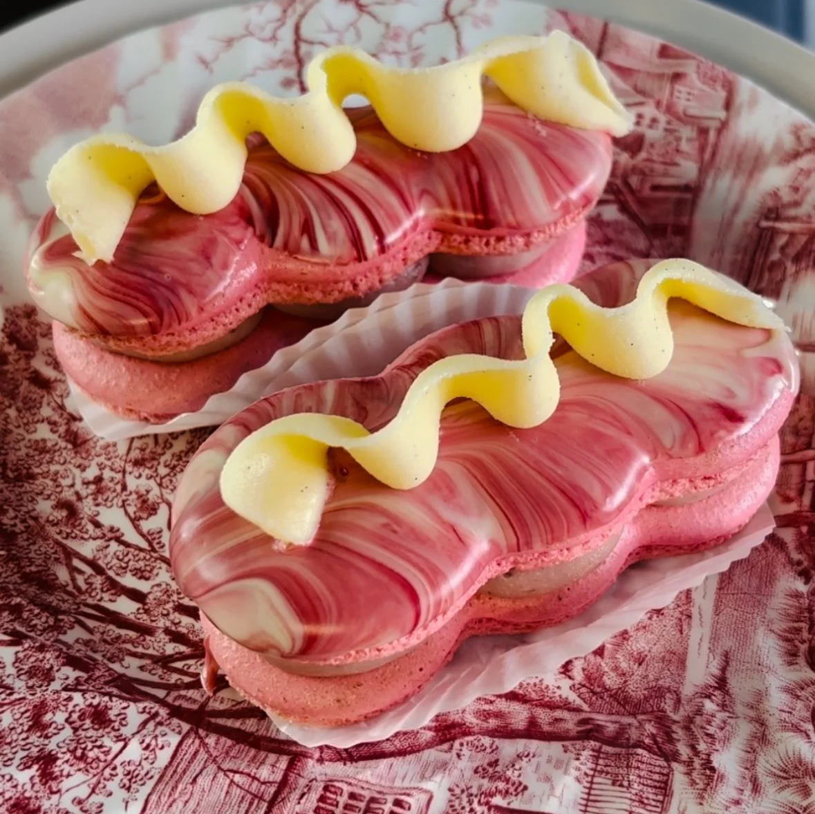 Dos pasteles decorativos de color rosa con ribetes blancos y trazo de chocolate, decorados con una espiral de glaseado blanco, en un plato decorativo rojo y blanco.