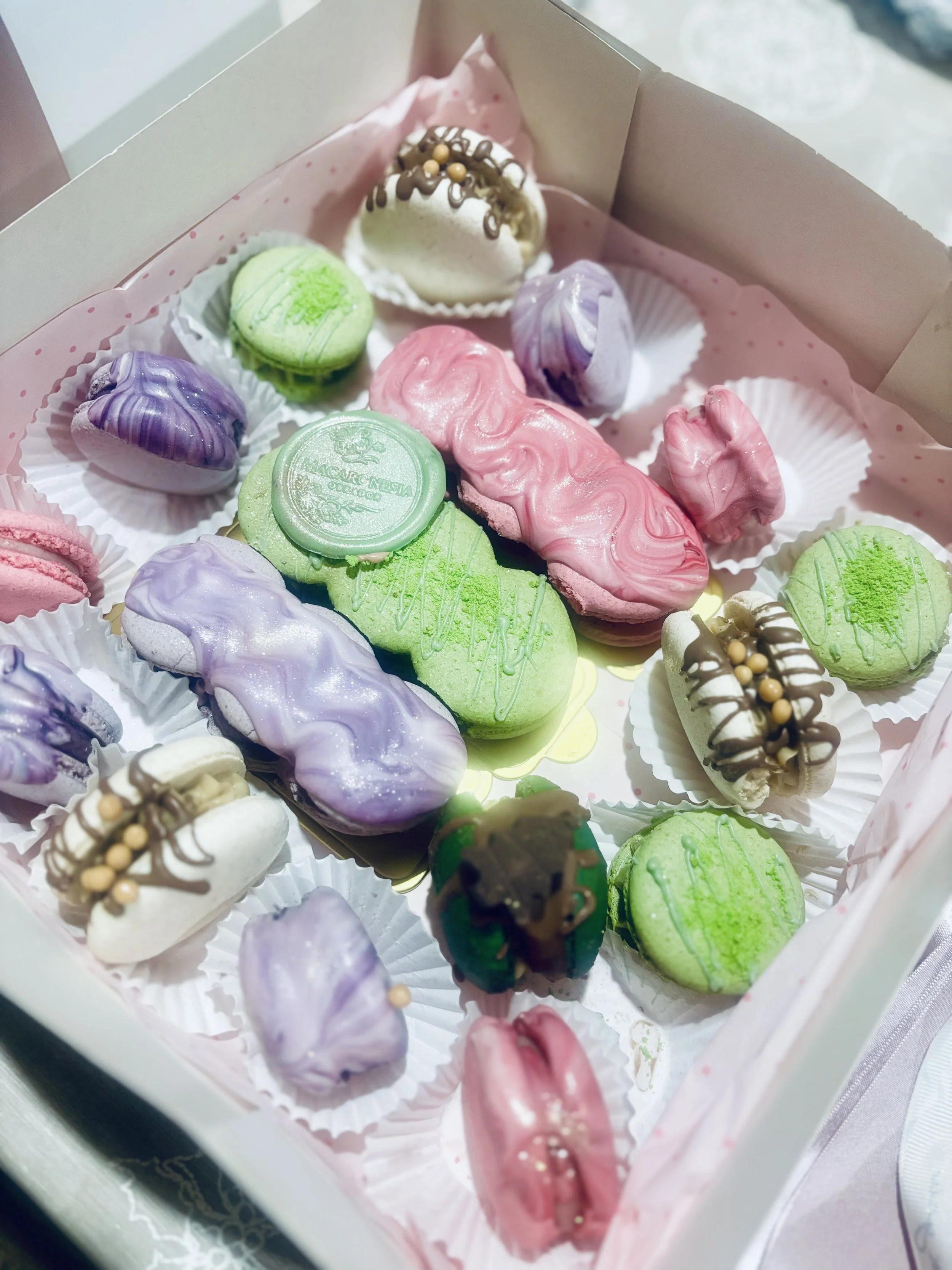 Caja con diversos macarons en colores morado, verde, rosa, y blanco decorados con chocolates y perlas comestibles.